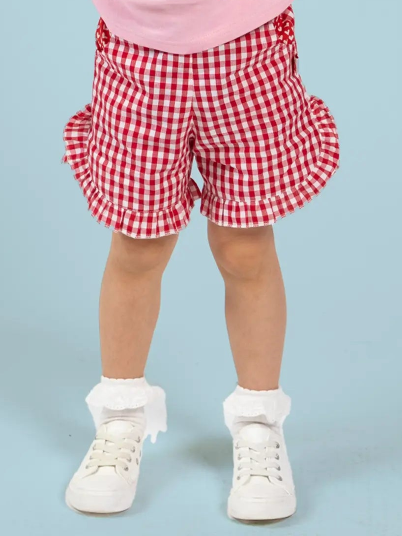 A child wearing Red Gingham Ruffle Shorts by Red Gingham, paired with white frilly socks and white sneakers, stands against a light blue background.