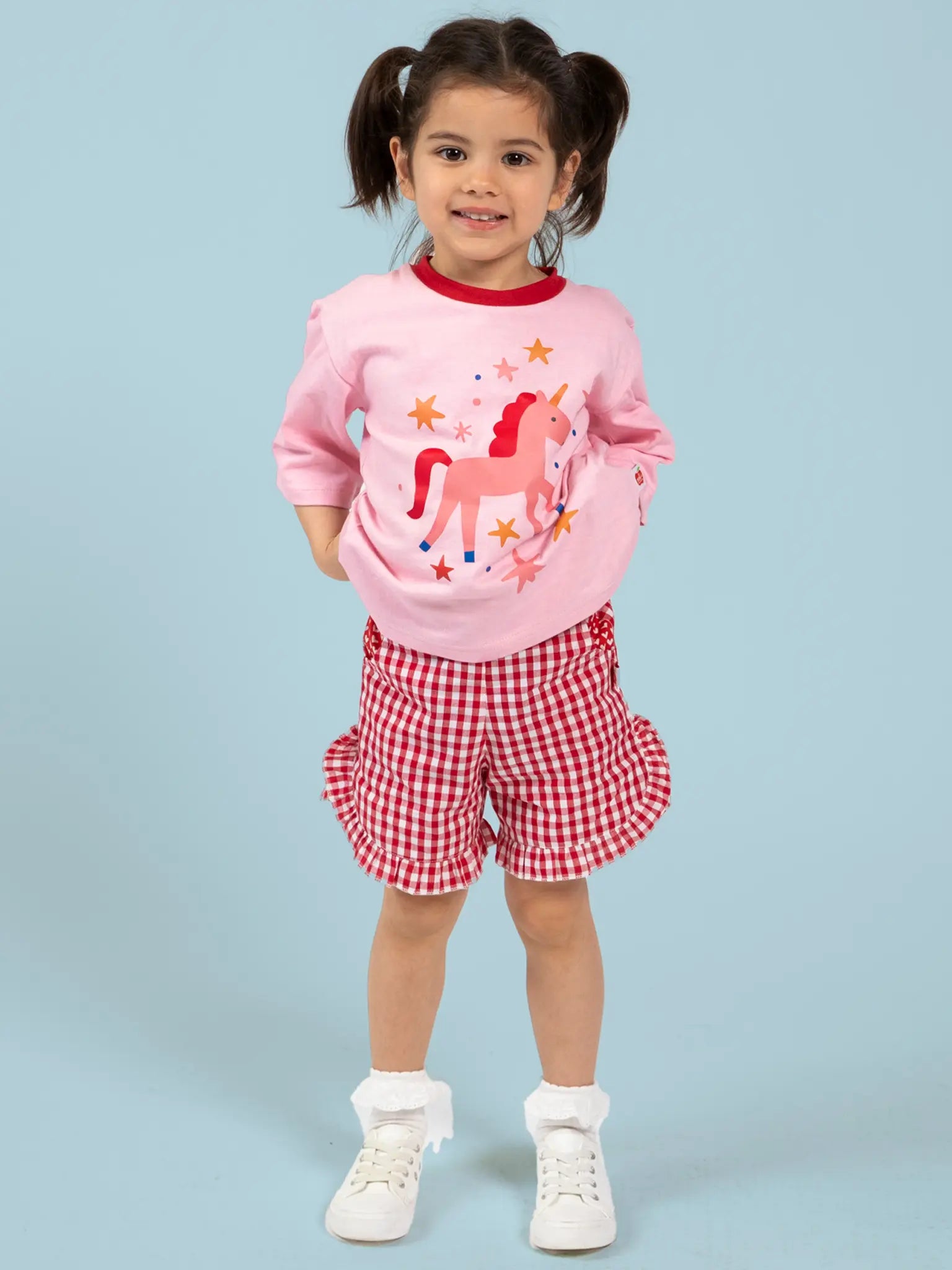 A young girl stands against a light blue background wearing Red Gingham Ruffle Shorts by Red Gingham, paired with a pink unicorn sweatshirt, white socks, and white shoes.