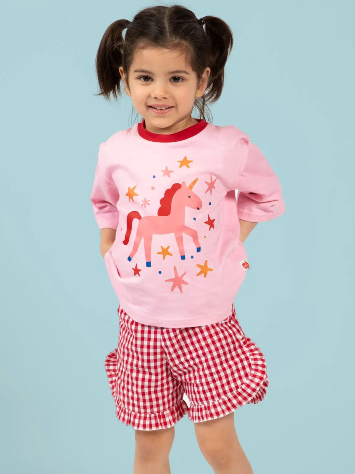 A young girl with pigtails stands against a light blue background, wearing Red Gingham Ruffle Shorts by Red Gingham and a pink unicorn shirt.