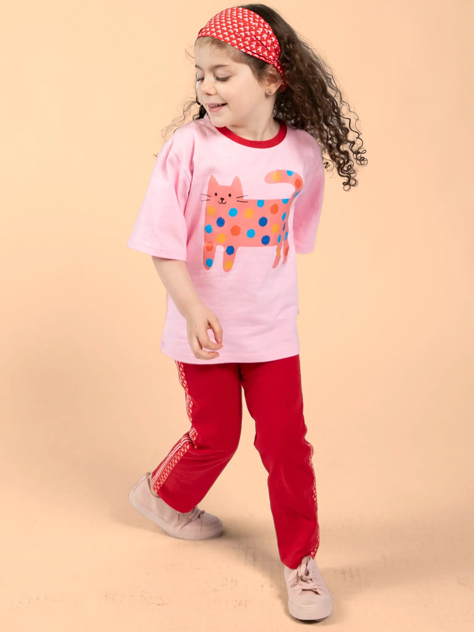 A young girl in 100% cotton sportswear—a pink cat-print t-shirt, Essentials Red Jersey 2 Stripe Pants, pink shoes, and a red polka-dot headband—stands against a plain beige background.