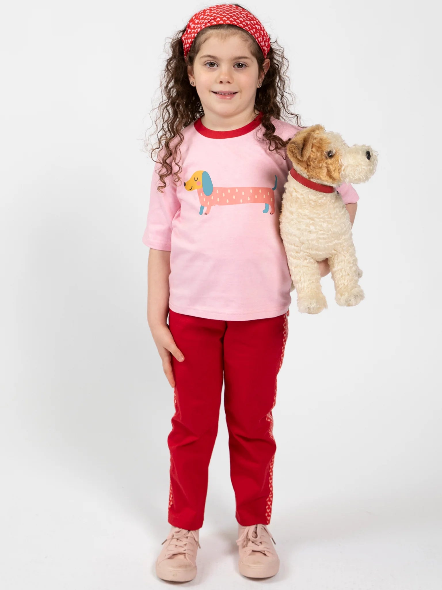 A young girl with curly hair in a pink dog-graphic shirt, Essentials Red Jersey 2 Stripe Pants, and a red headband holds a stuffed dog toy while standing against a plain background.