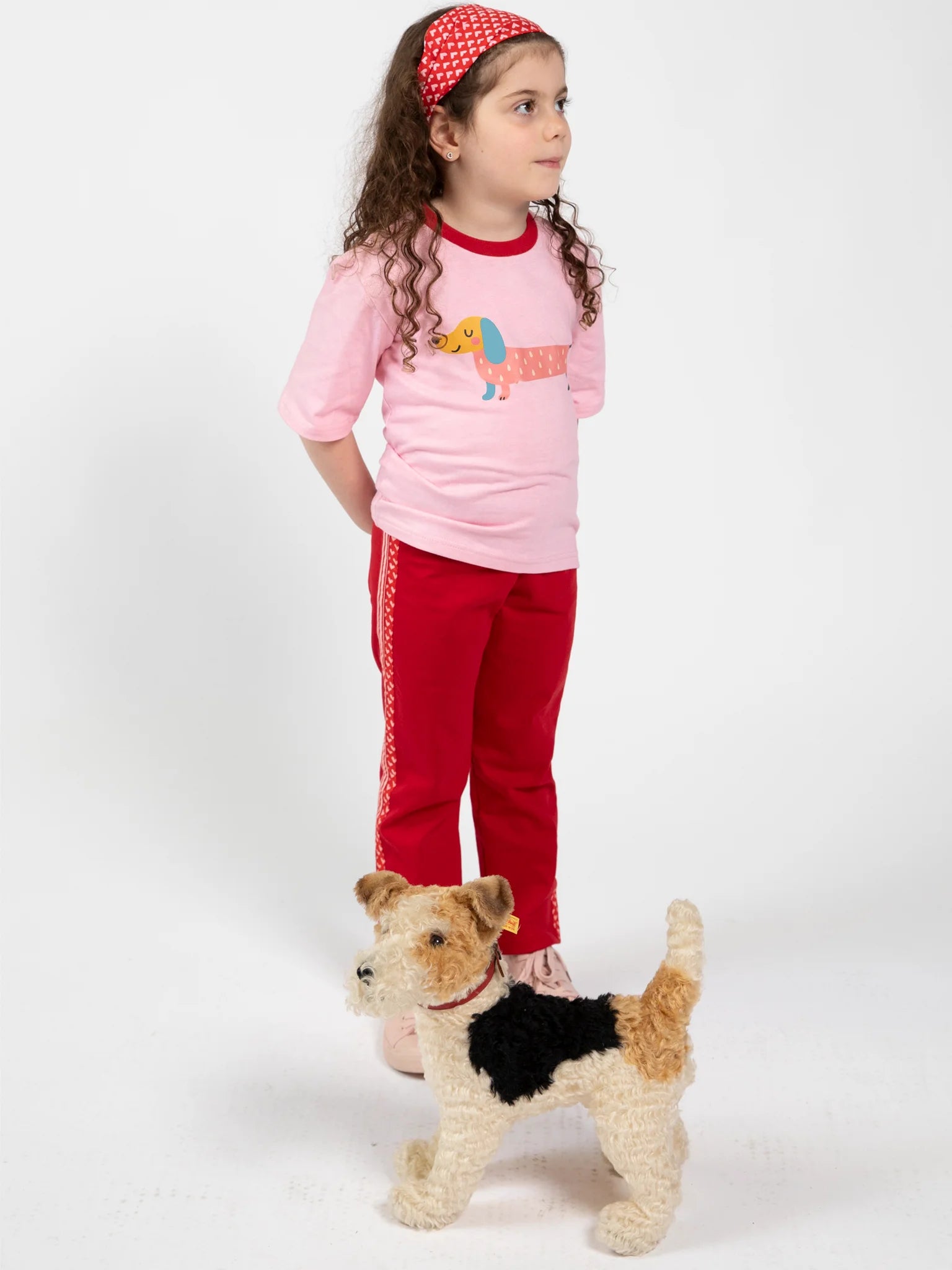 A young girl in Essentials sportswear—a pink dachshund graphic shirt and Red Jersey 2 Stripe Pants—stands by a small tri-color dog against a plain white background.