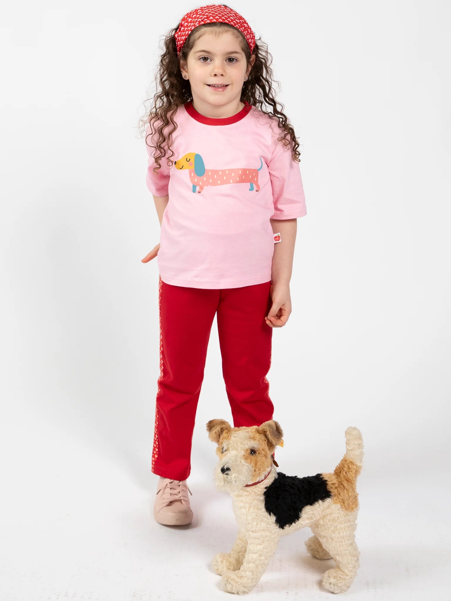 A young girl wears Essentials Red Jersey 2 Stripe Pants, a pink dachshund t-shirt, and a red headband as she stands next to a small plush dog against a plain white background.