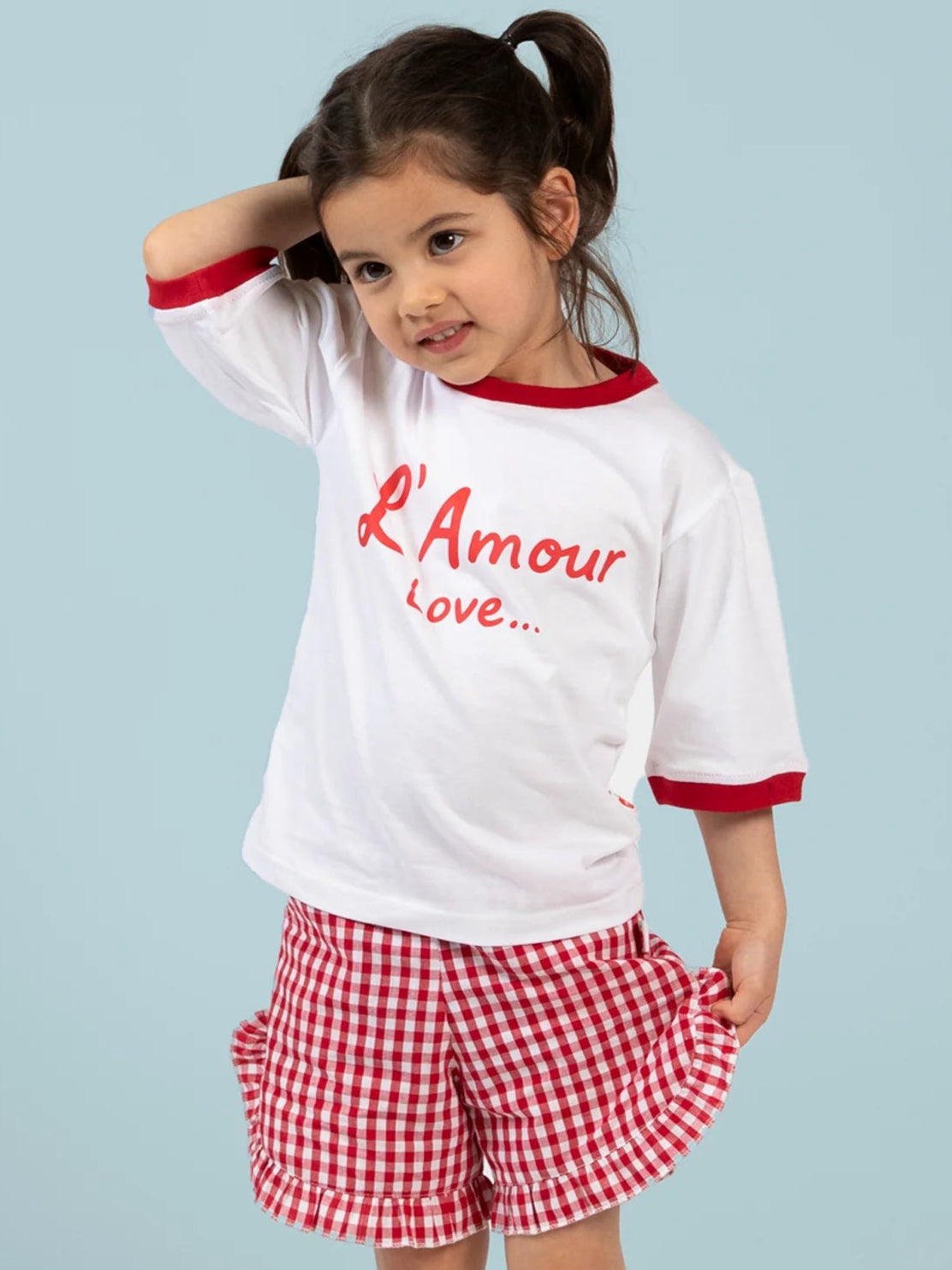 A young girl wears Red Gingham brand ruffle shorts with a red gingham pattern and an elastic waist, paired with a white T-shirt that has "L'Amour Love..." in red, standing against a light blue background.