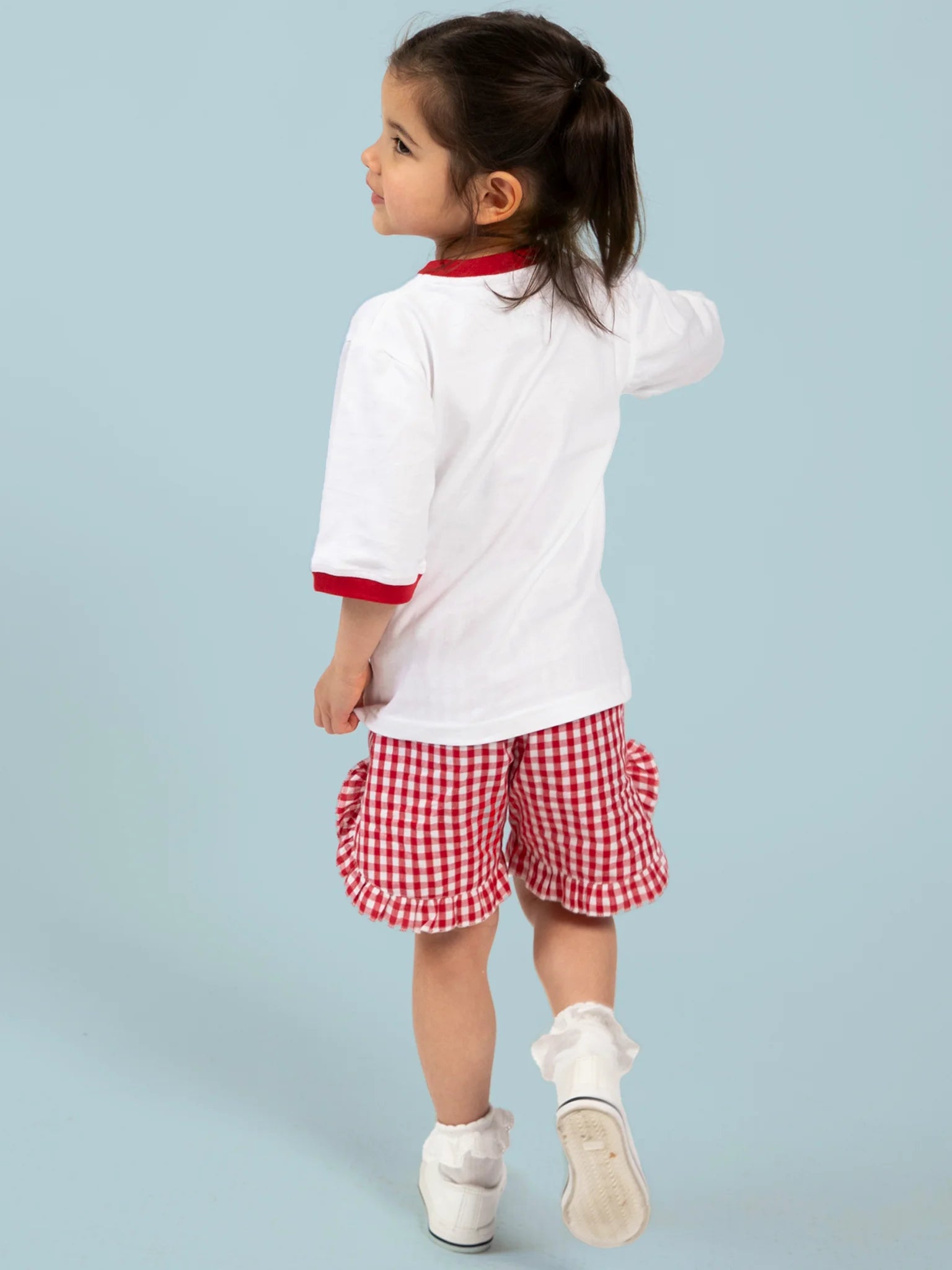 A young child with dark hair in a ponytail wears the Essentials White Cherry Tee with Red Binding, standing and facing away against a light blue background. The hand-printed Cherry Bow print is made in Australia.