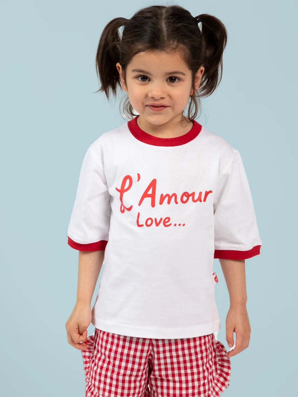 A young child with dark hair in pigtails wears the Essentials L'Amour (Love) Tee with Red Binding, made from 100% cotton, paired with red and white checkered shorts, standing against a light blue background.