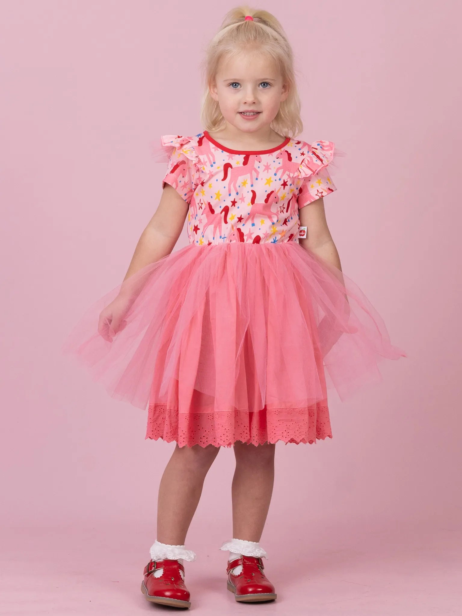 A young girl with blonde hair in a ponytail stands against a solid pink studio background, wearing a pink and red floral print dress with a pink tulle tutu skirt and red Mary Jane shoes.