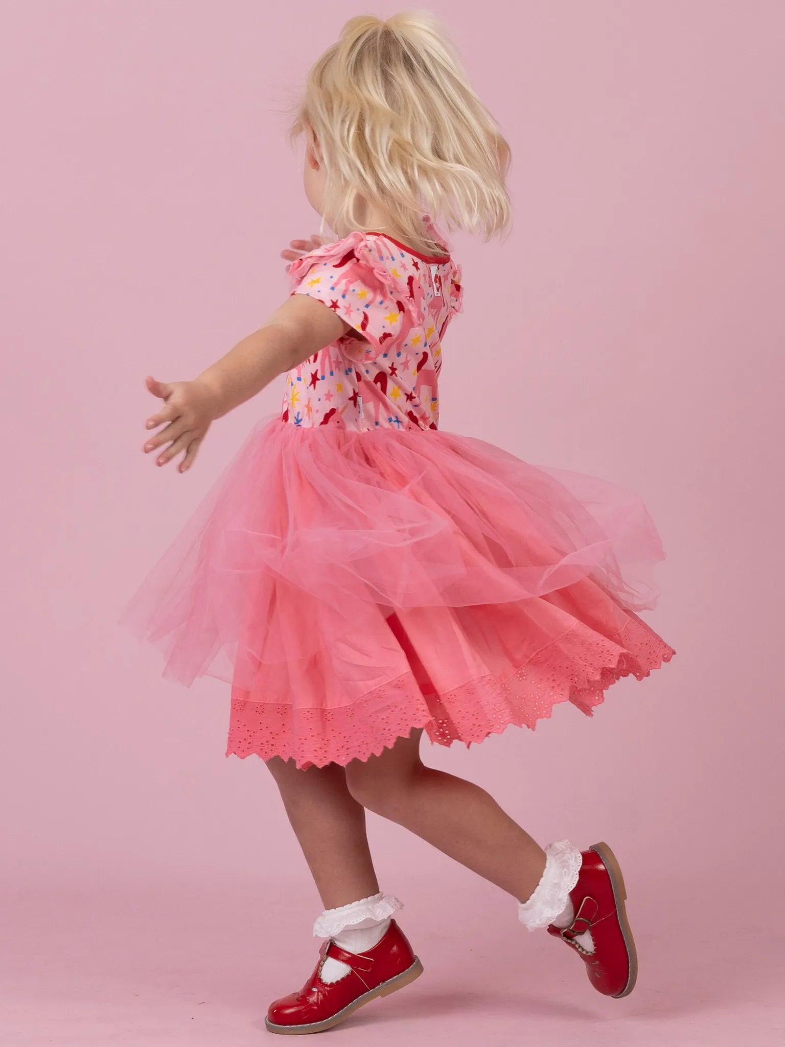 A young child wearing a pink tulle tutu dress with a colorful floral bodice and red shoes, photographed against a solid pink studio background.