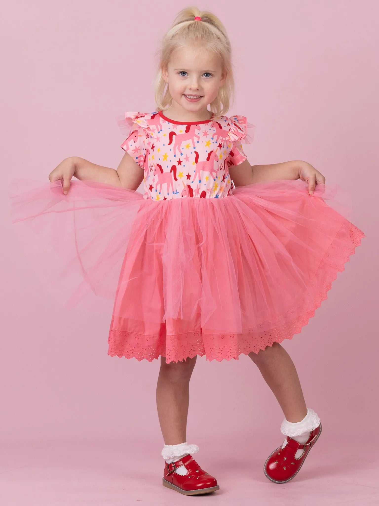A young blonde girl wearing a pink dress with a colorful ice cream pattern bodice and a fluffy pink tulle skirt, posing against a solid pink background.