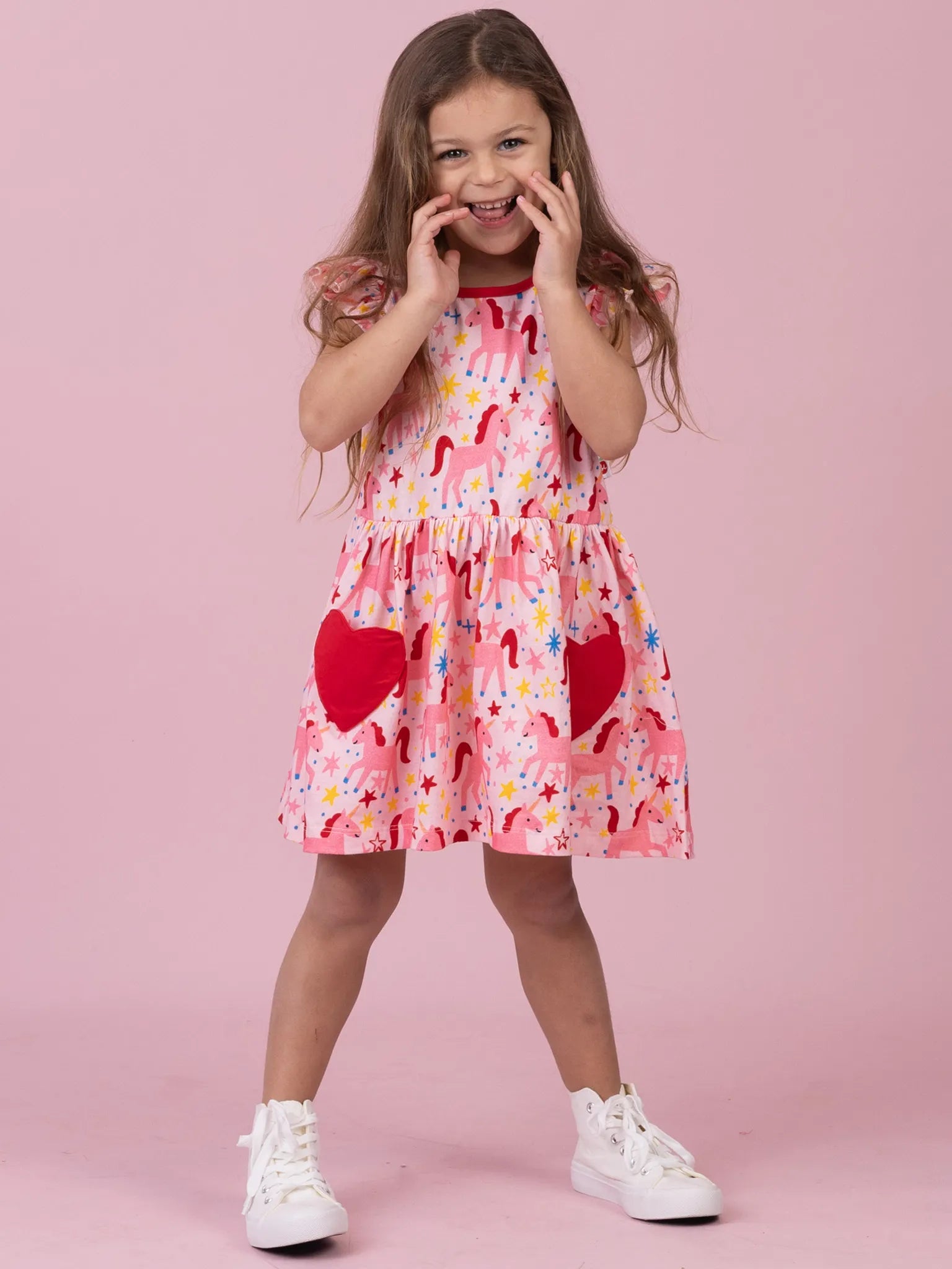 A young girl with long brown hair stands against a solid pink studio background wearing a tiered pink dress with a red and multicolor print, featuring two large red heart pockets, and white sneakers.