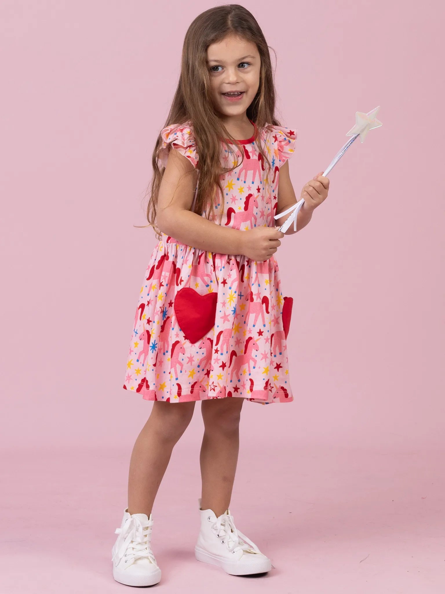 A young girl with long brown hair stands against a solid pink studio background, wearing a pink dress with a rainbow and heart pattern, holding a white star wand, and wearing white sneakers.