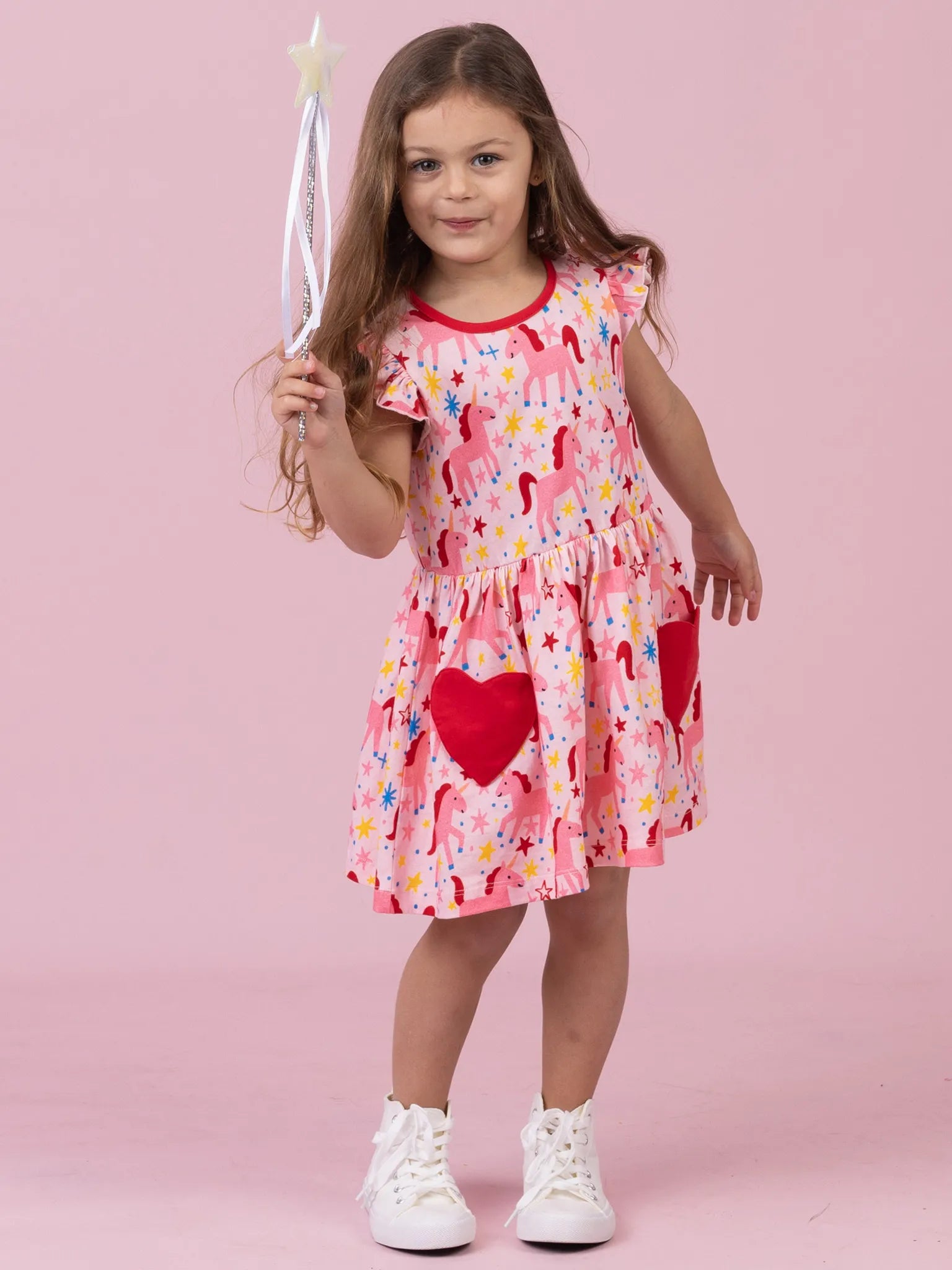 A young girl stands against a solid pink studio background wearing a pink ruffled dress with a colorful unicorn and heart pattern, holding a white star wand, and wearing white sneakers.