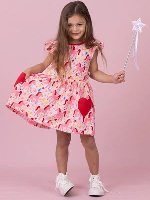 A young girl with long brown hair stands against a solid pink studio background, wearing a pink dress with a red, yellow, and blue unicorn print, red heart-shaped pockets, and holding a silver star wand.
