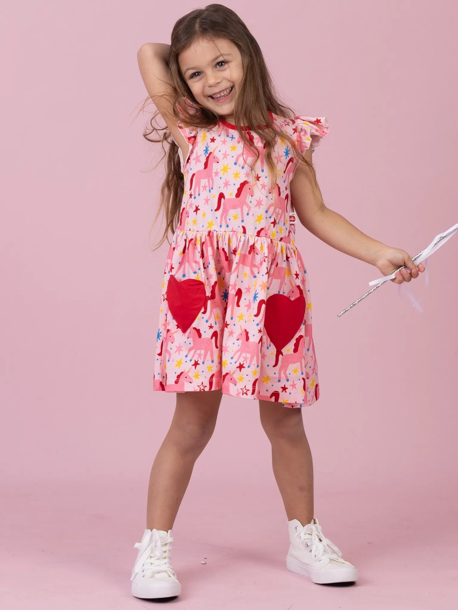 A young girl with long brown hair stands against a solid pink studio background, wearing a pink tiered dress with a white flamingo print, red heart patches on the skirt, and white sneakers. She is holding a silver toy sword.