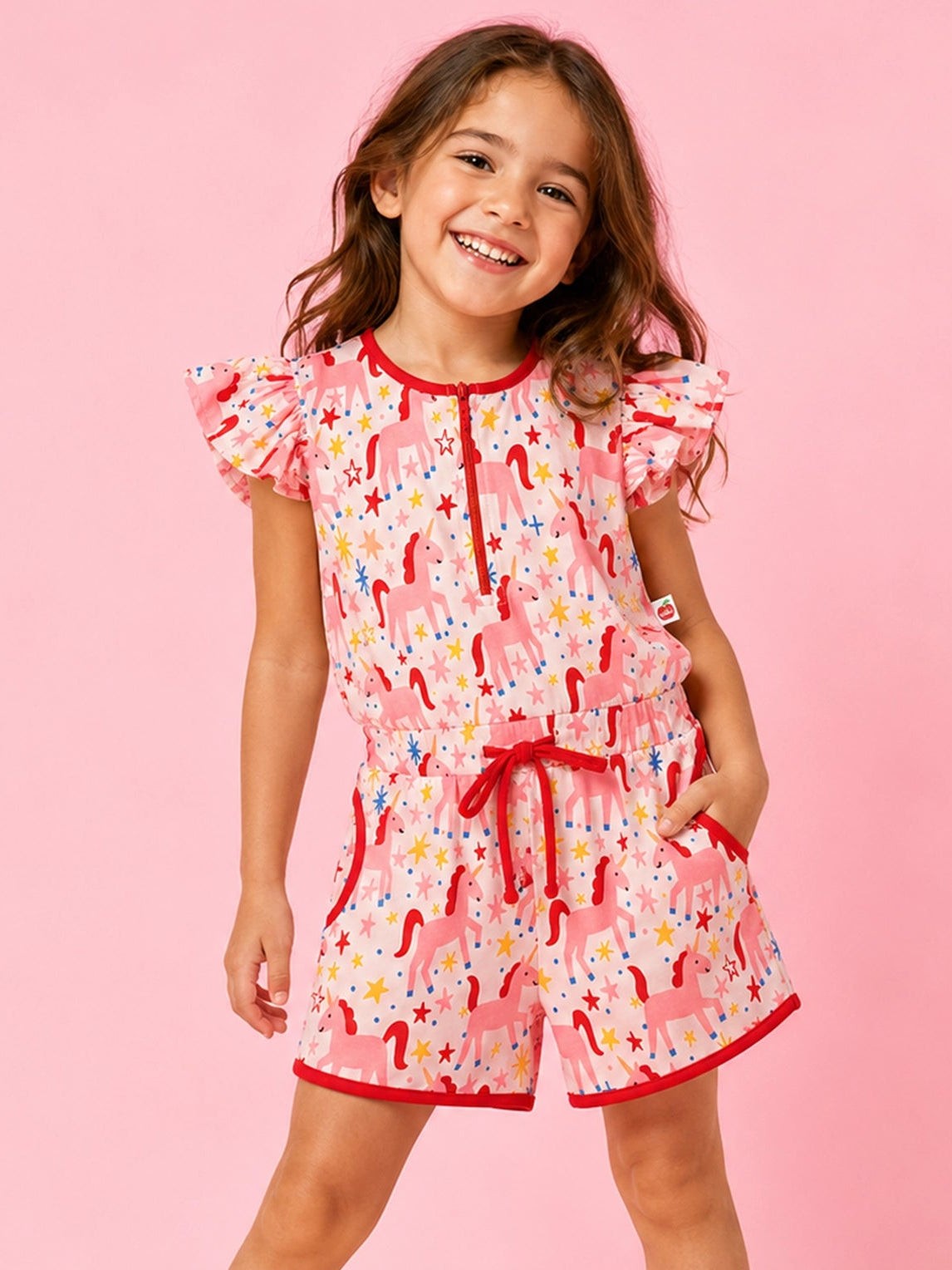 A young girl modeling a pink and white unicorn-themed romper with ruffled sleeves and a matching pink shoe, set against a solid pink studio background.