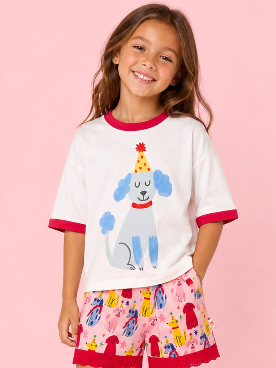 A young girl stands against a solid pink background wearing a white ringer T-shirt with red trim and a blue poodle graphic, paired with pink patterned shorts featuring red ruffle hems and white sneakers.