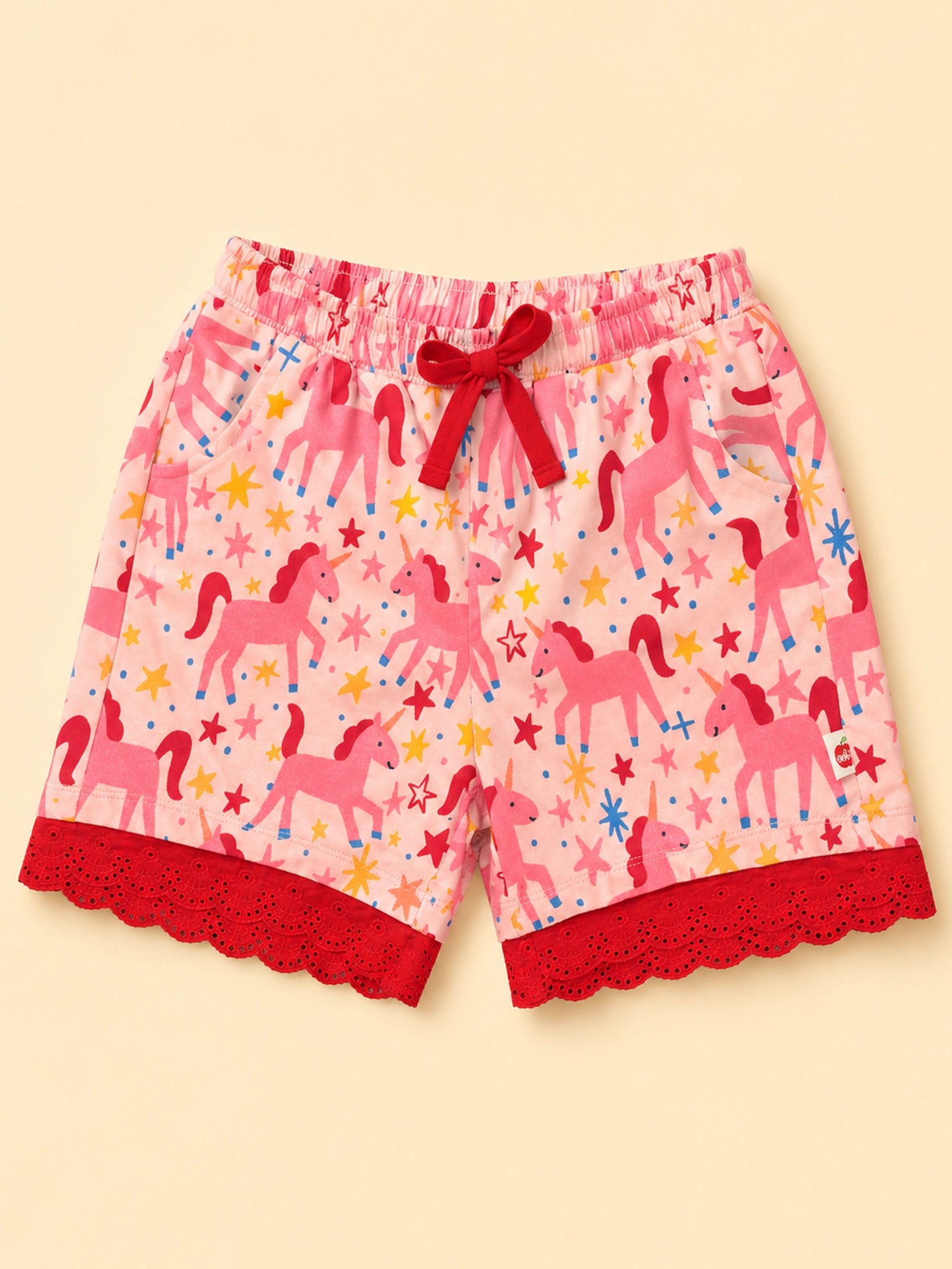 Pink children's shorts with an allover unicorn and star print, featuring a red drawstring bow at the elastic waistband and red scalloped lace trim at the hem, shown against a pale yellow background.