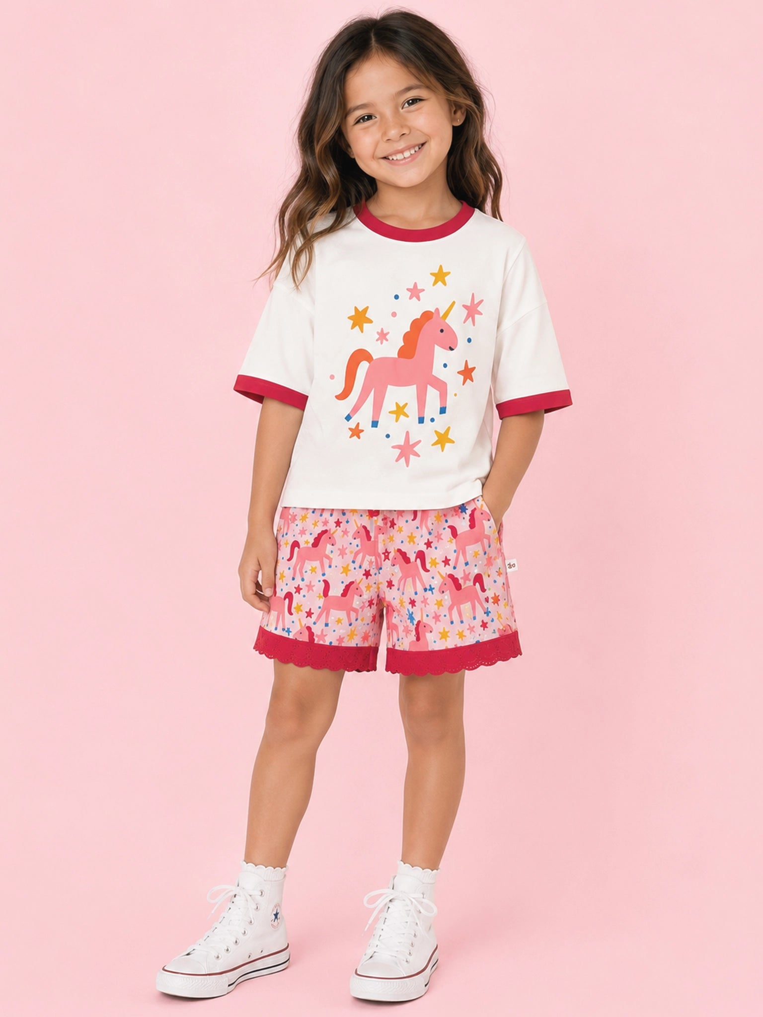 A young girl stands against a solid pink studio background wearing a matching ringer t-shirt and shorts set. The white t-shirt features a pink unicorn graphic with yellow stars and red collar/cuff trim, while the pink shorts have a multicolor unicorn print and red ruffled hems.