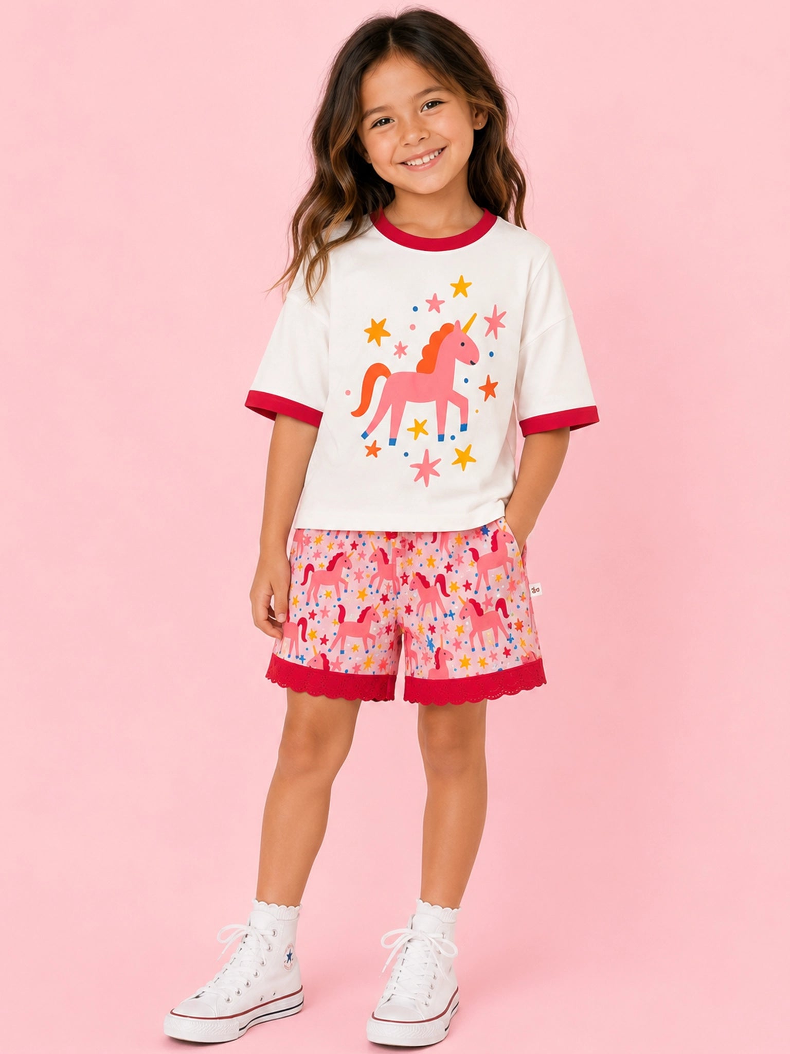A young girl stands against a solid pink studio background wearing a matching two-piece set. The set features a white short-sleeve ringer T-shirt with red trim and a pink unicorn graphic, paired with matching pink patterned shorts with red ringer trim.
