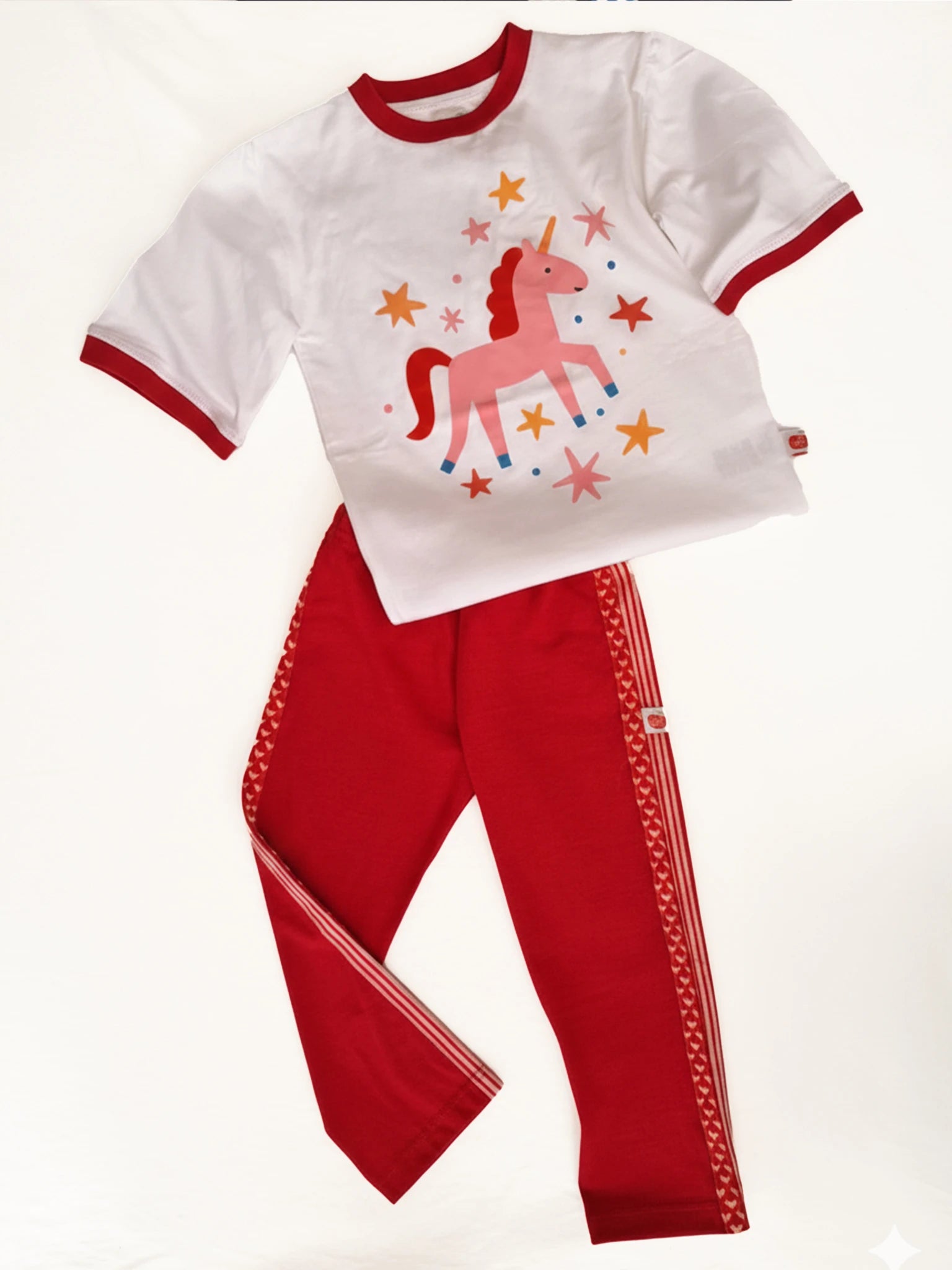 The Essentials girls sportswear set includes a 100% cotton white t-shirt with a pink unicorn and stars, paired with Red Jersey 2 Stripe Pants featuring heart patterns.