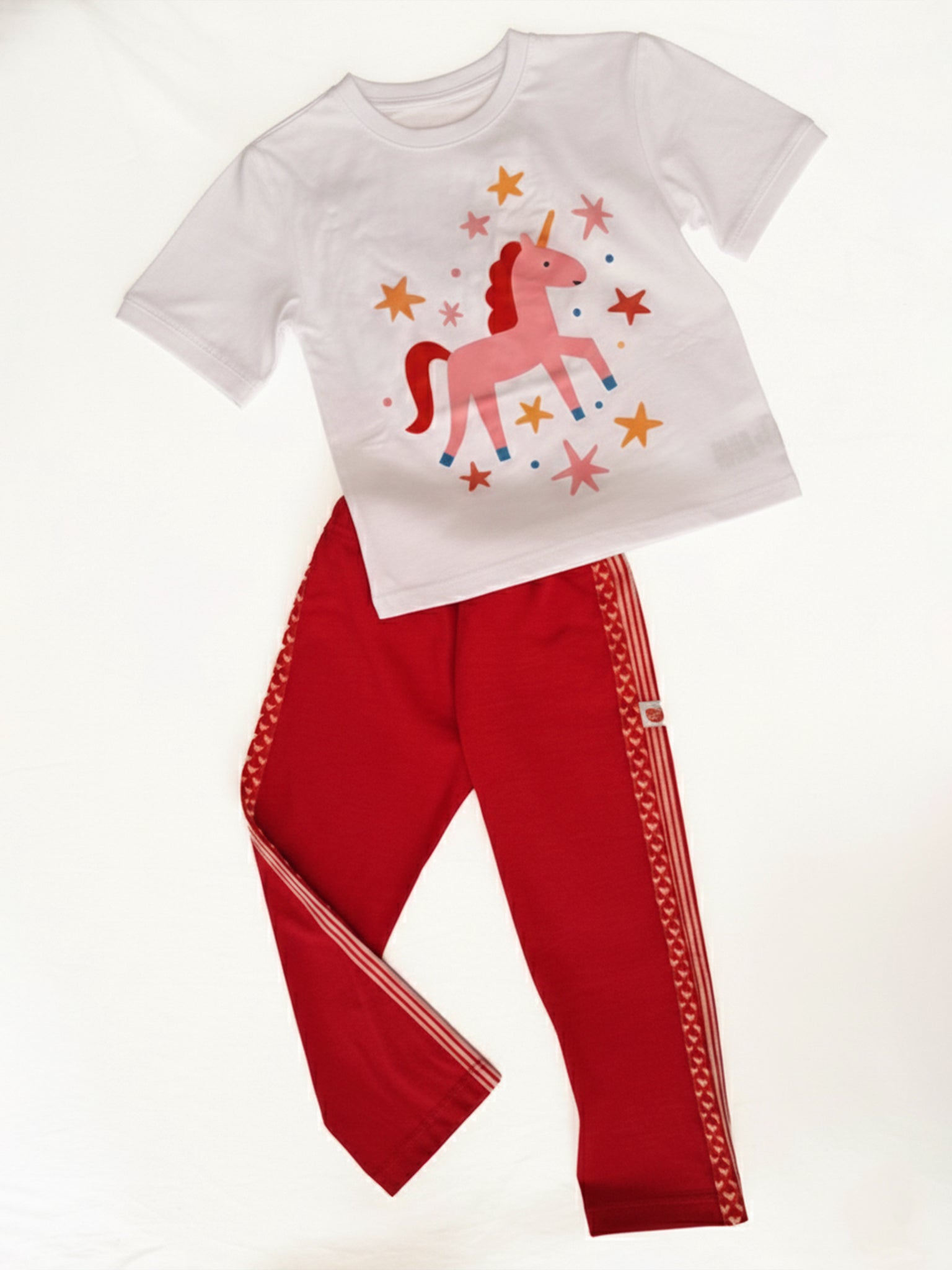 The Essentials Unicorn Printed Tee—a white oversized cotton top with a pink unicorn and stars graphic—is paired with red pants featuring white side stripes and a patterned trim. Personalisation available, sizes 2Y to Women.
