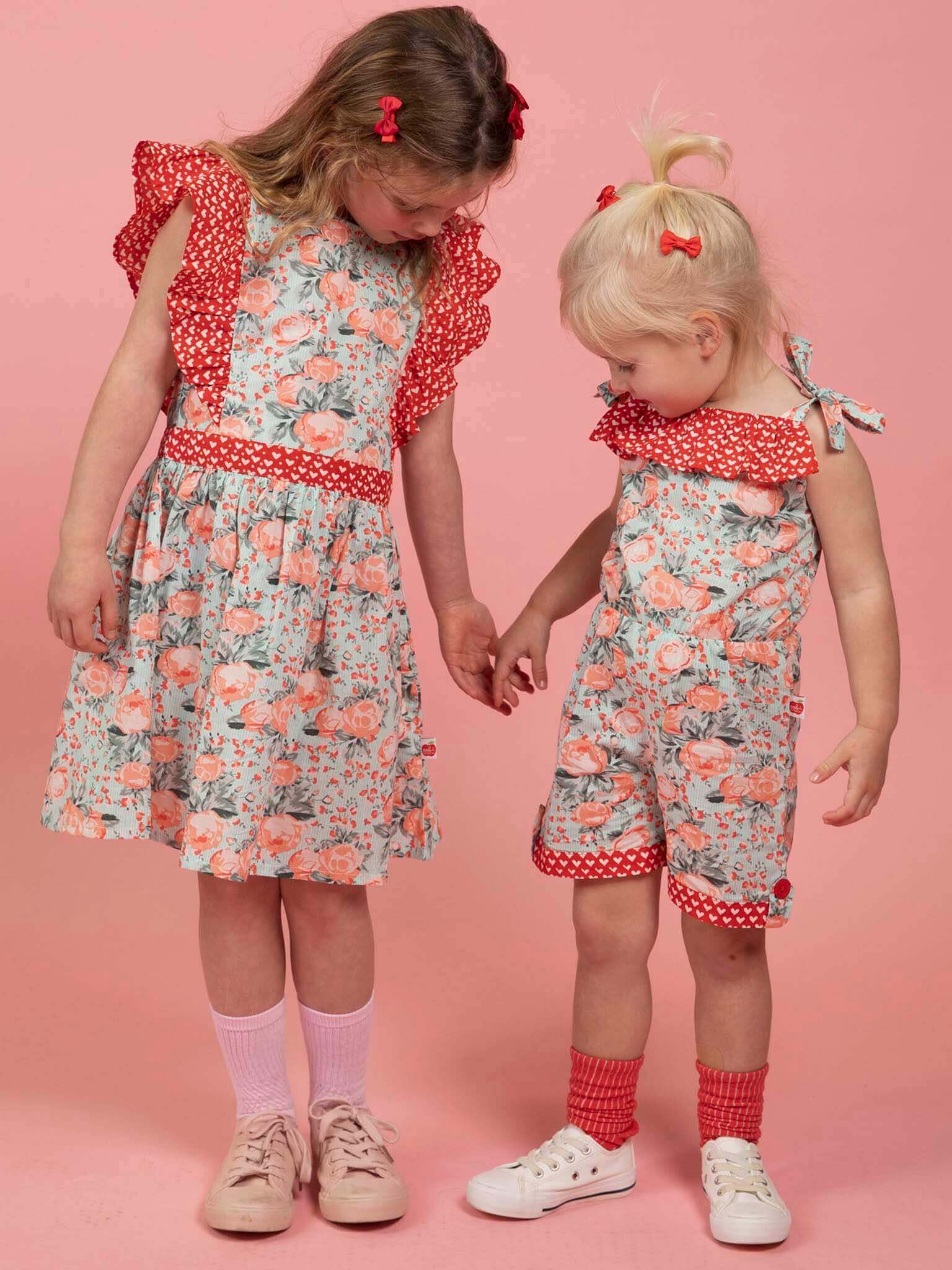 Two young girls wearing Vintage Roses Jayne Dresses by Vintage Roses, featuring coordinated floral designs, stand on a pink background holding hands and looking down.