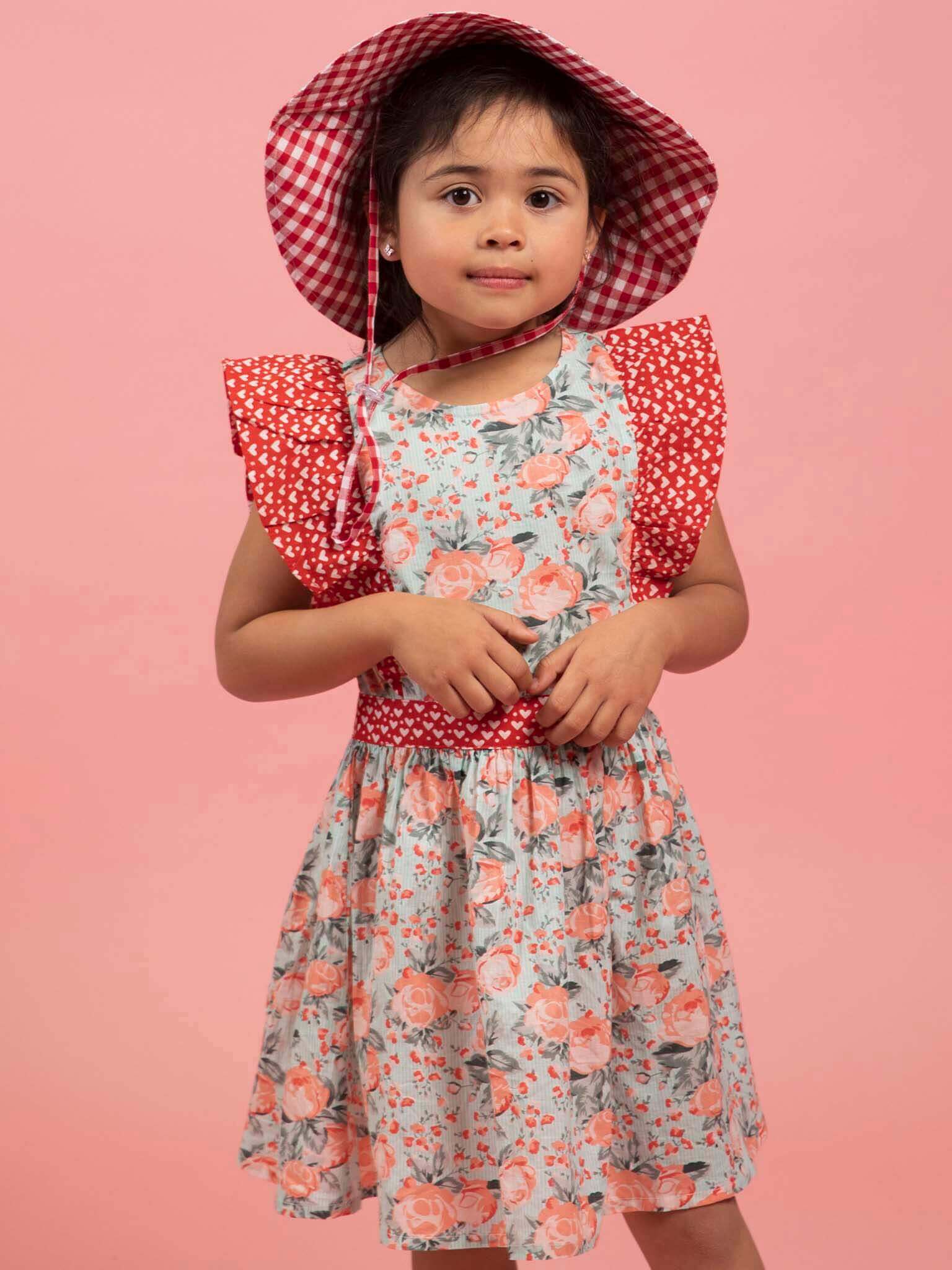 Child wearing a floral dress with red accents and a matching hat on a pink background