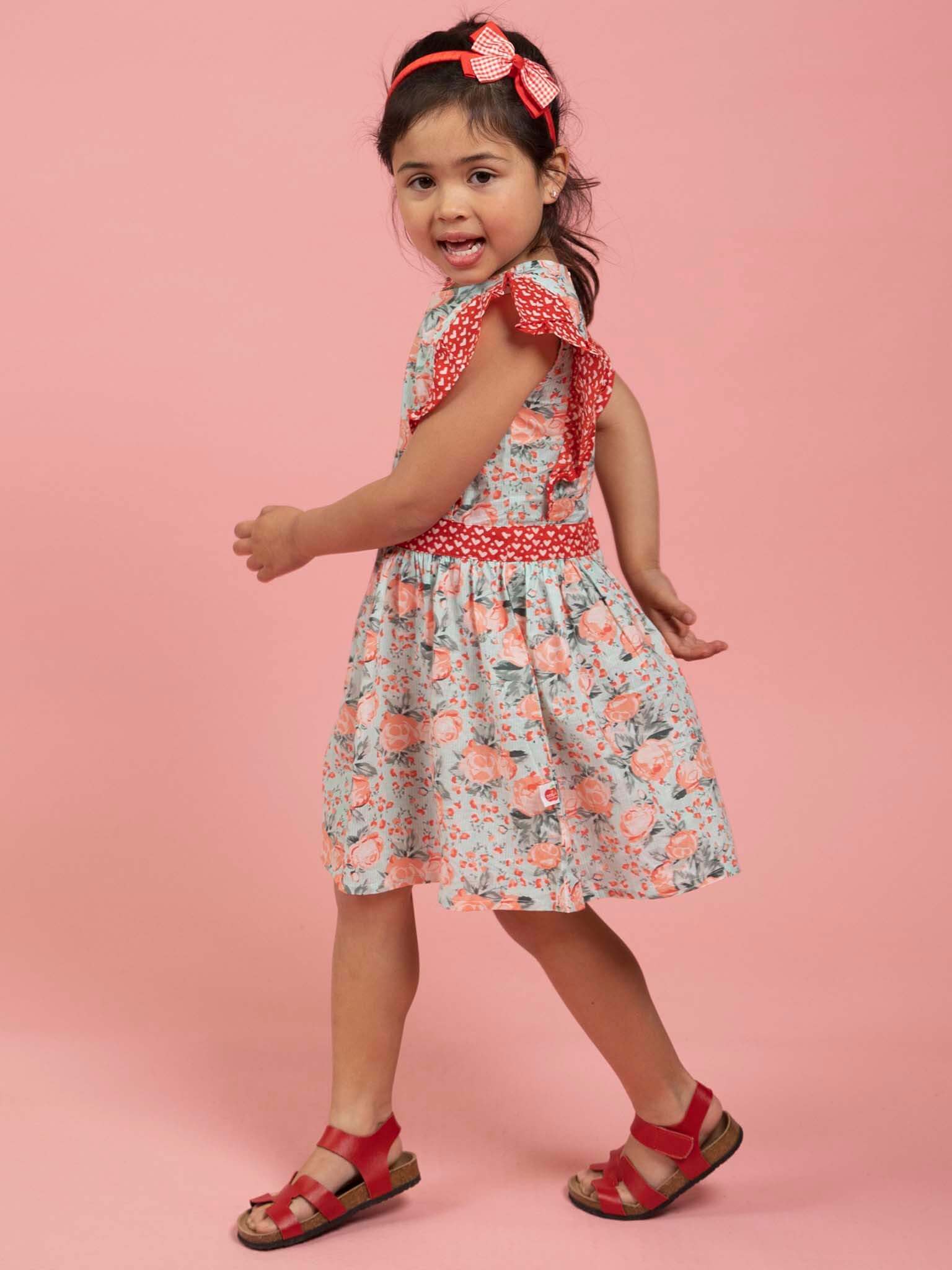 A young girl in the Vintage Roses Jayne Dress with floral patterns and red accents, paired with red sandals, stands against a pink background, looking back over her shoulder.