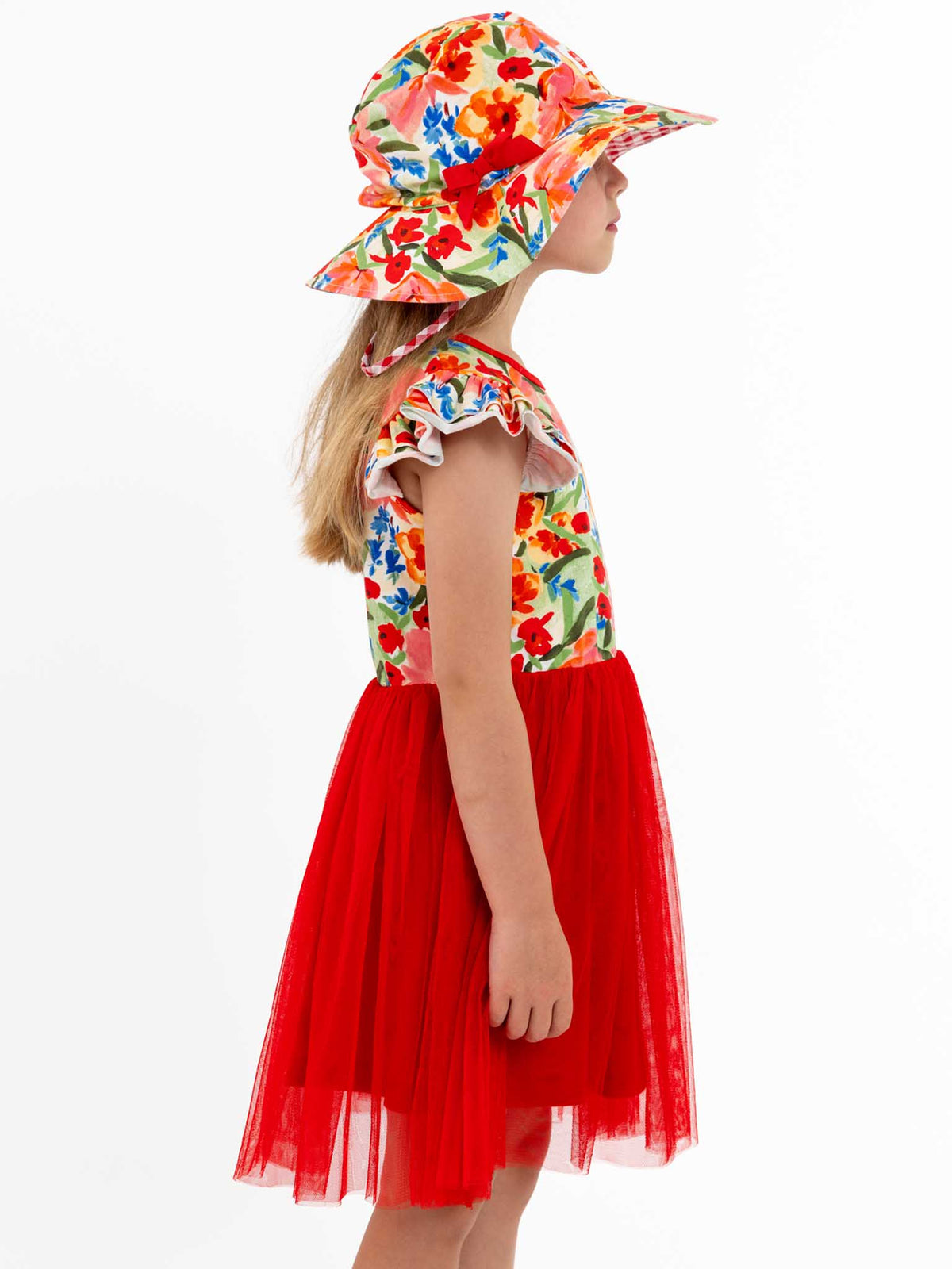 A young girl in a Wild Roses Chloe Ponytail Hat and a floral dress with a red tulle skirt stands in profile against a white background.