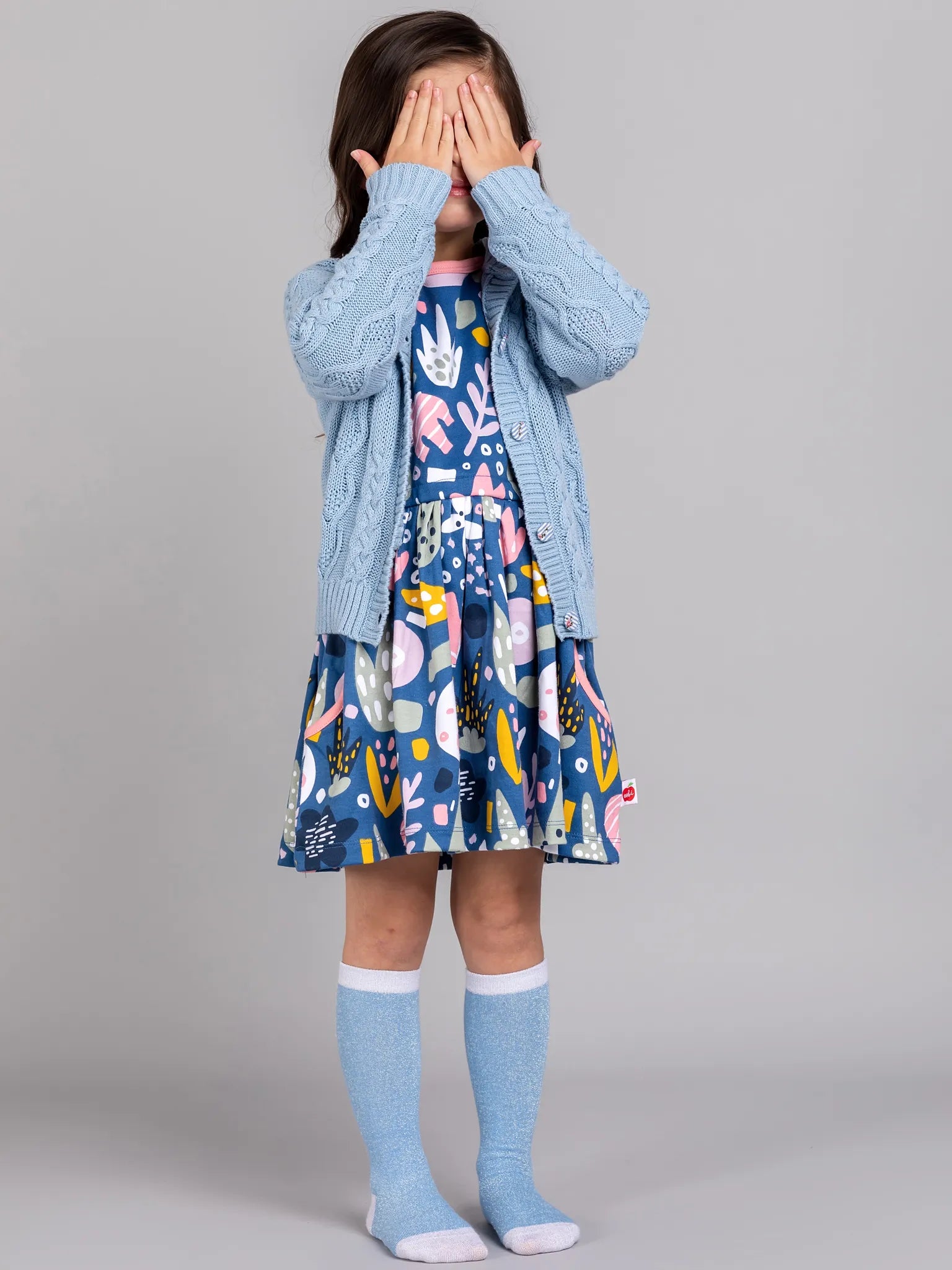 Child wearing a blue dress with colorful patterns and a matching cardigan against a gray background
