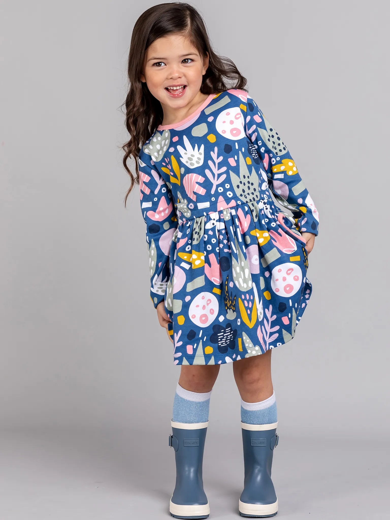 Child wearing a blue dress with colorful patterns and blue rain boots on a gray background