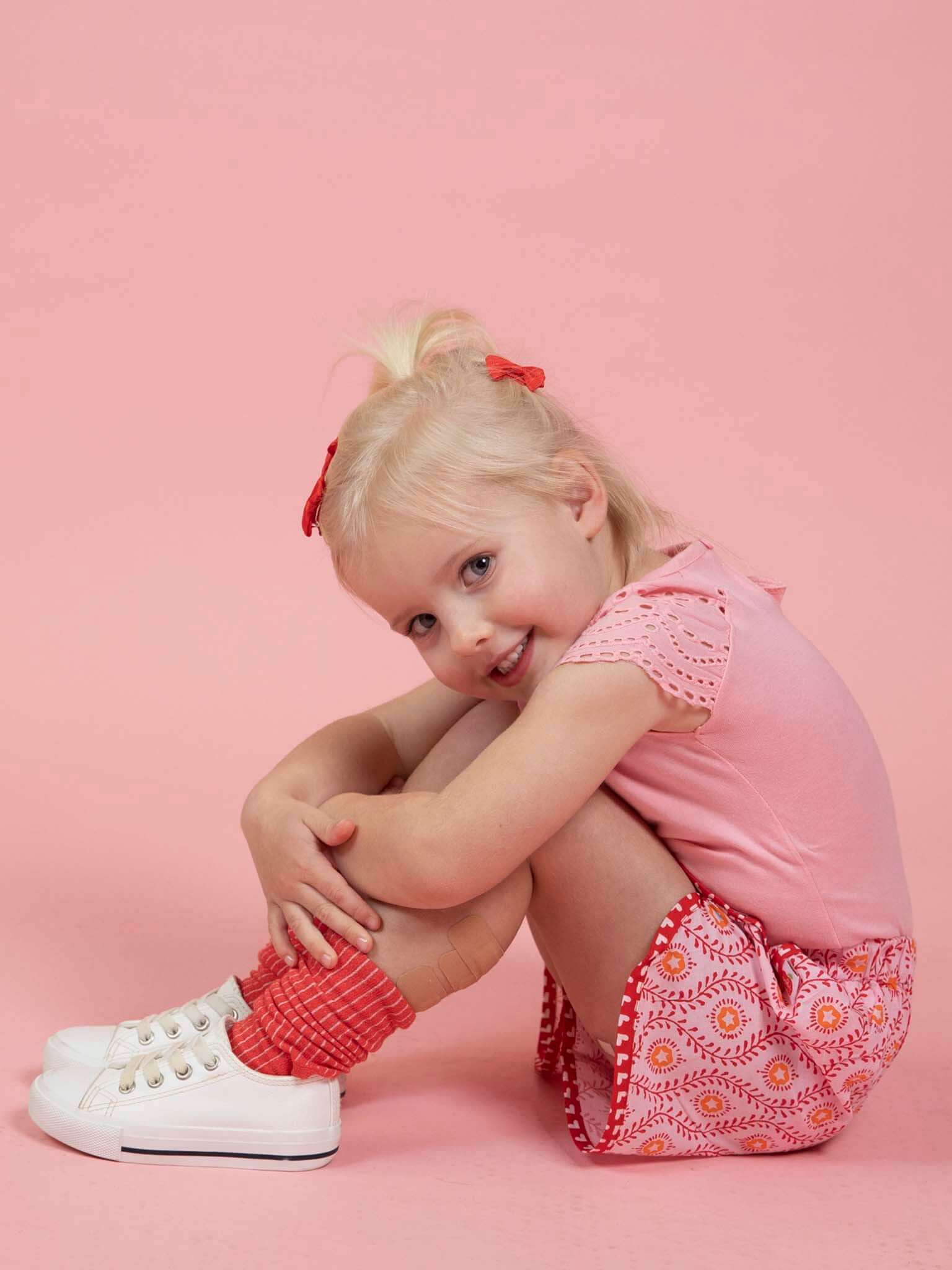 A young girl with blonde hair sits on the floor against a pink background, smiling at the camera while wearing a pink top, red socks, white sneakers, and Woodblock Flower Curved Shorts from Woodblock Flower.