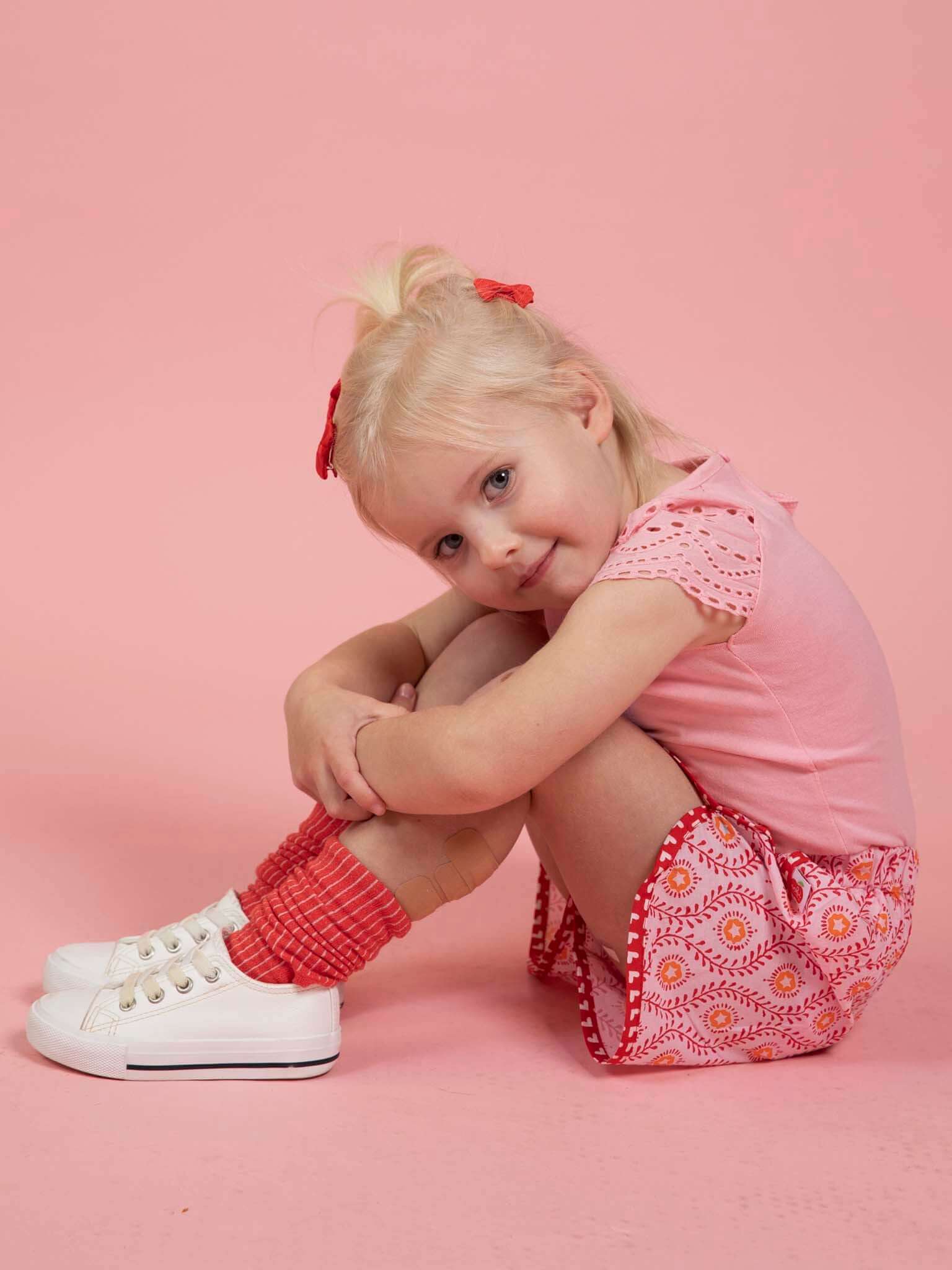 A young child with blonde hair, wearing a pink shirt, Woodblock Flower Curved Shorts by Woodblock Flower, red socks, and white shoes sits curled up on a pink floor against a pink background, smiling at the camera.