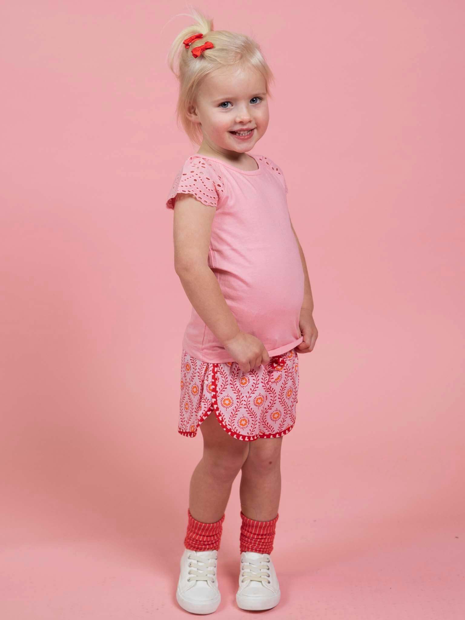 A young child with blonde hair in a ponytail, wearing a pink shirt, Woodblock Flower Curved Shorts by Woodblock Flower, red socks, and white shoes stands smiling against a pink background.