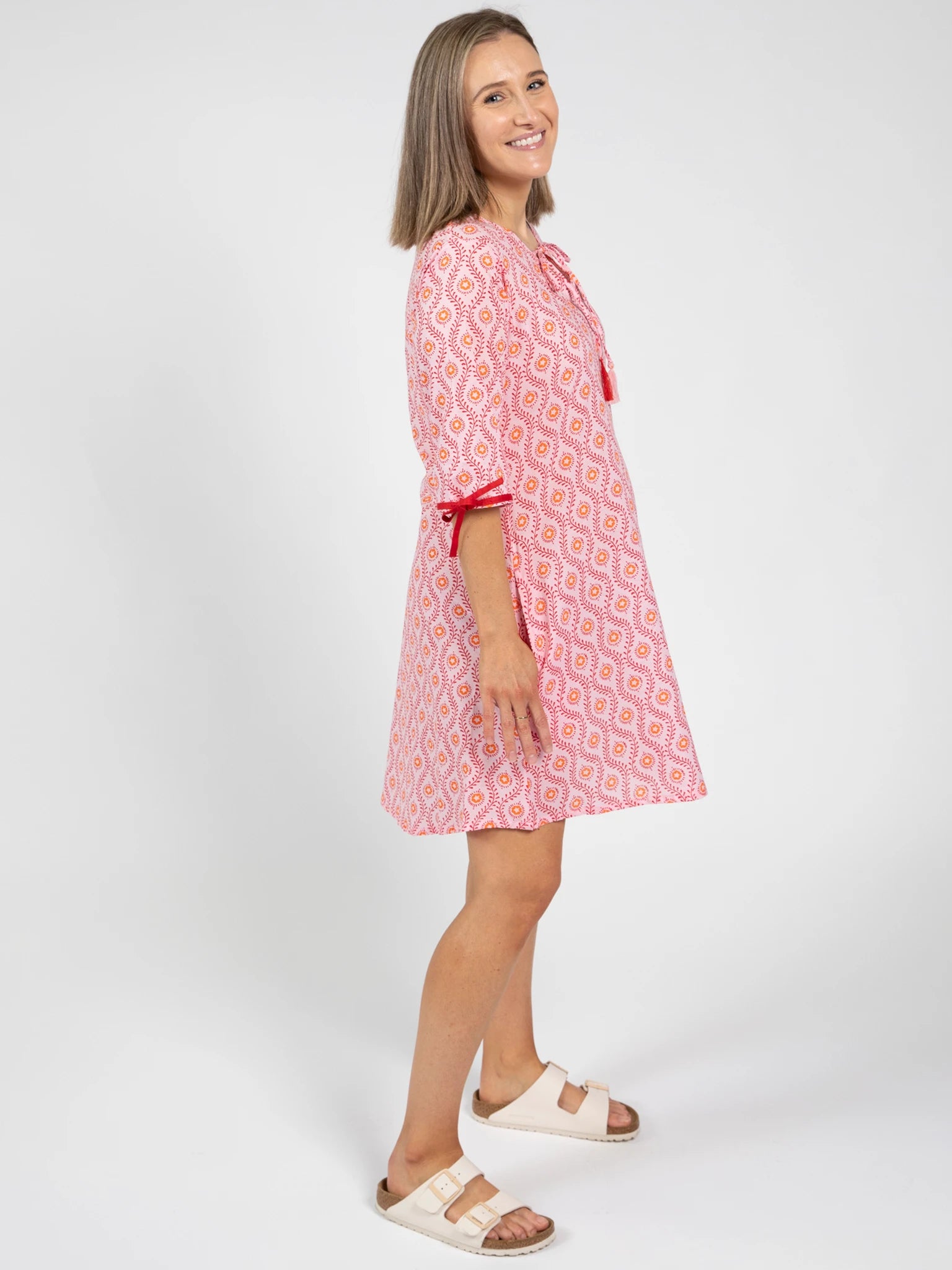 A woman stands sideways, smiling, in the Woodblock Women's Resort Dress by Woodblock Flower—a pink and white woodblock print, knee-length kaftan with three-quarter sleeves and white sandals—set against a plain white background.