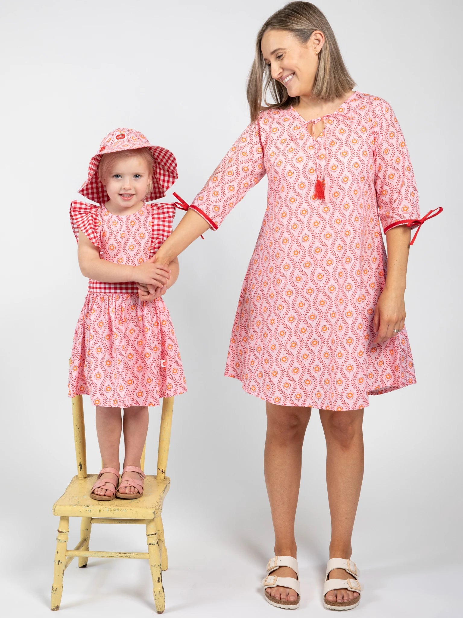 A woman in a Woodblock Flower Woodblock Women's Resort Dress stands beside a smiling girl in a matching dress and hat, who is standing on a yellow chair.