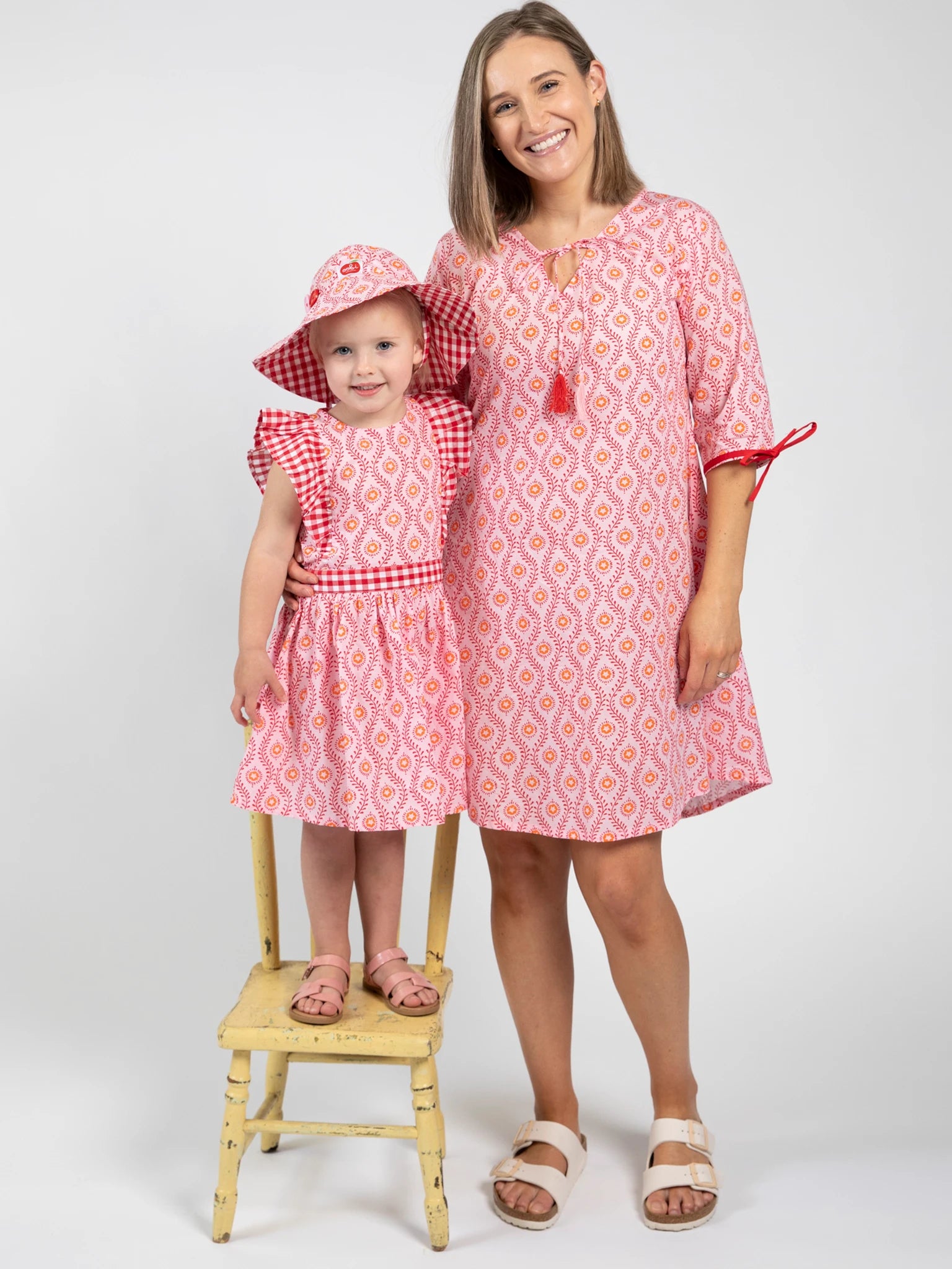 An adult and a child wear matching Woodblock Flower Jayne Dresses in pink; the child stands on a small yellow chair, also wearing a coordinating summer hat.