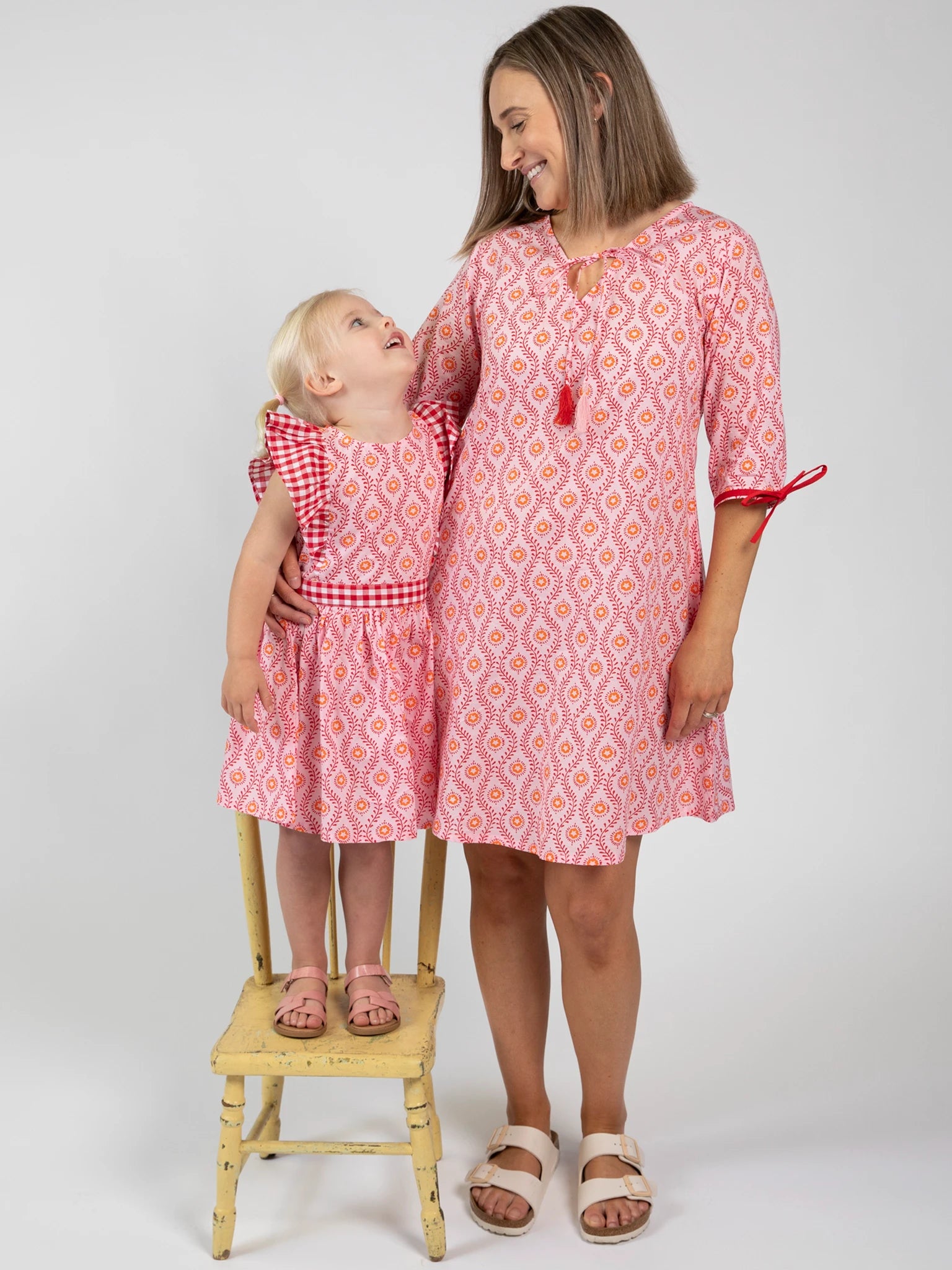 A woman and a child wear matching Woodblock Women's Resort Dress by Woodblock Flower; the child stands on a wooden chair, both smiling at each other.