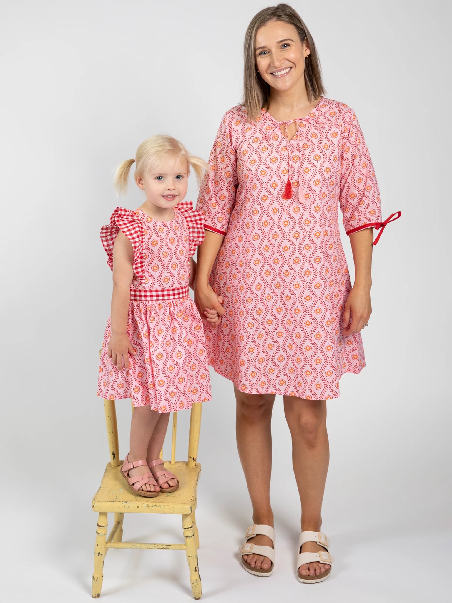 An adult woman and a young girl, both in matching pink Woodblock Flower Woodblock Women's Resort Dresses, stand together against a plain background; the woman stands on the floor while the girl stands on a yellow chair.