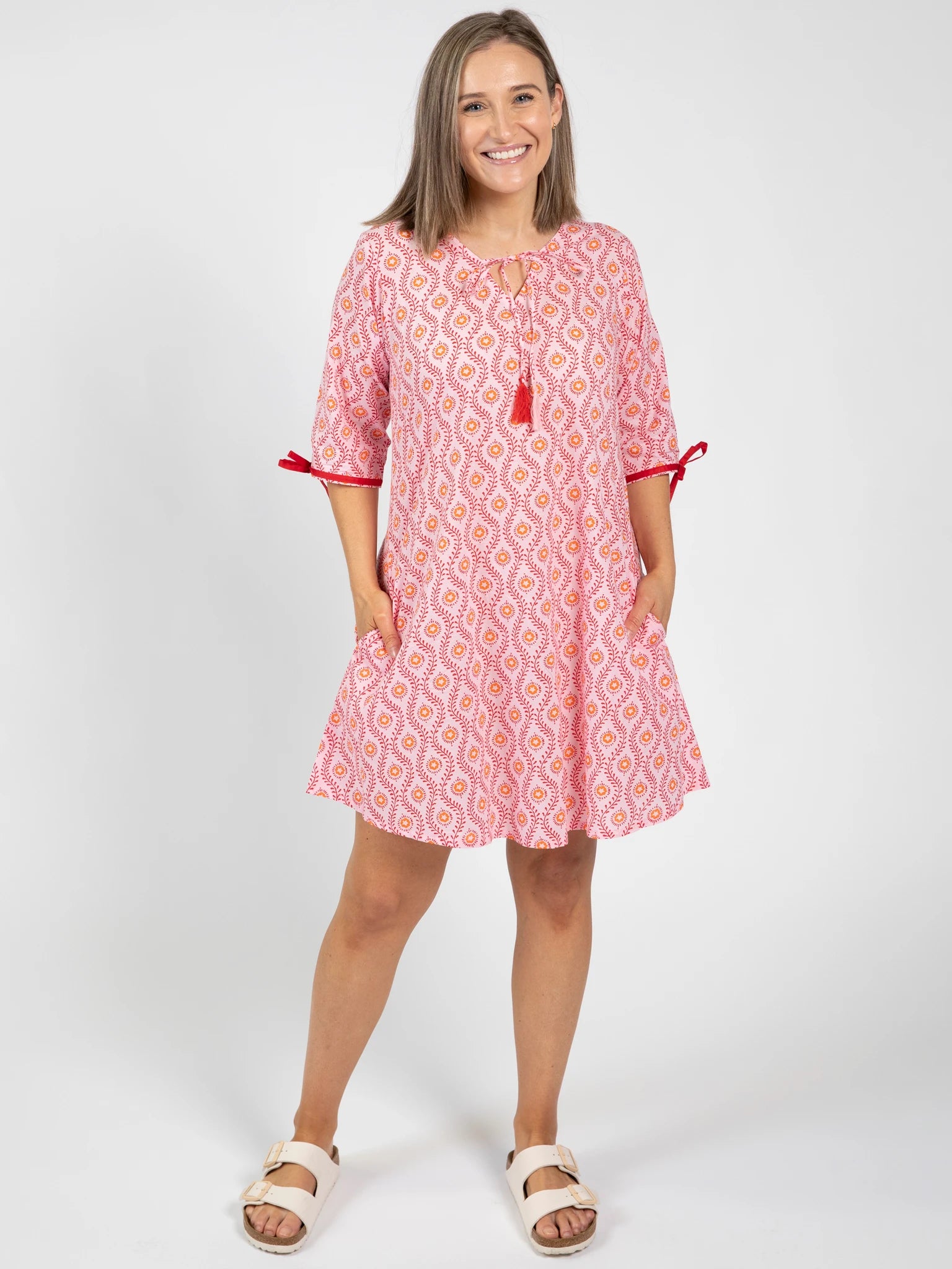 A woman stands smiling in the Woodblock Women's Resort Dress by Woodblock Flower—a knee-length pink and white woodblock print kaftan with red tassel and bow details—paired with white sandals against a plain white background.