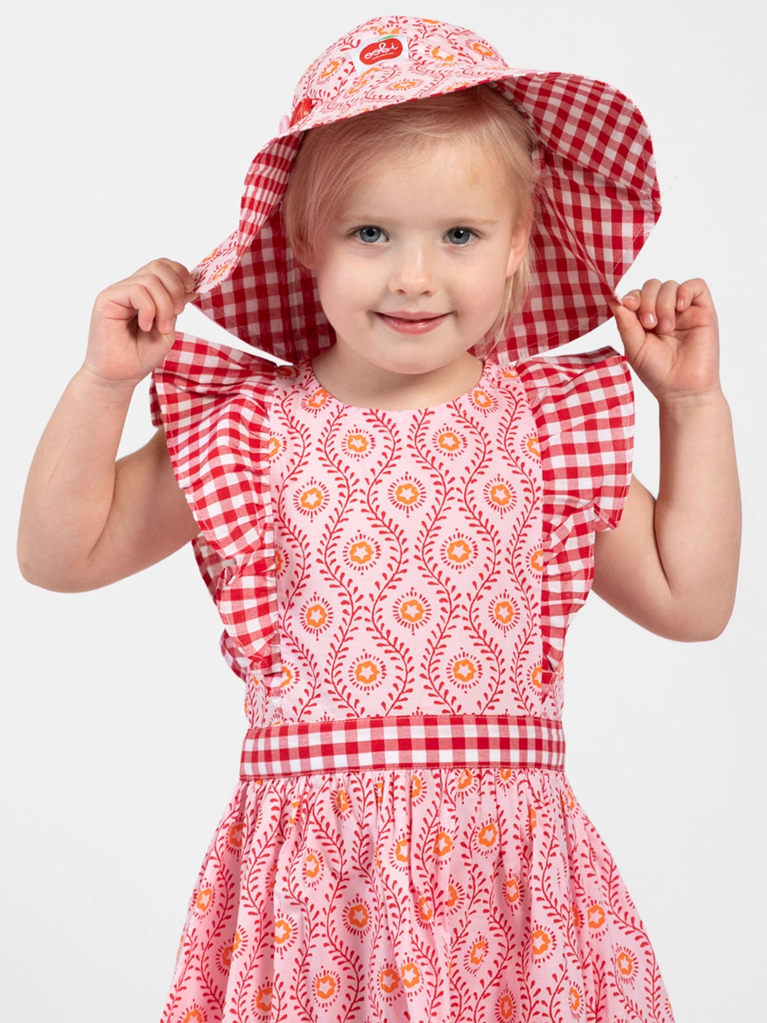 A young child smiles at the camera in the Woodblock Flower Jayne Dress by Woodblock Flower, paired with a matching wide-brimmed gingham hat, holding the hat with both hands against a plain background.