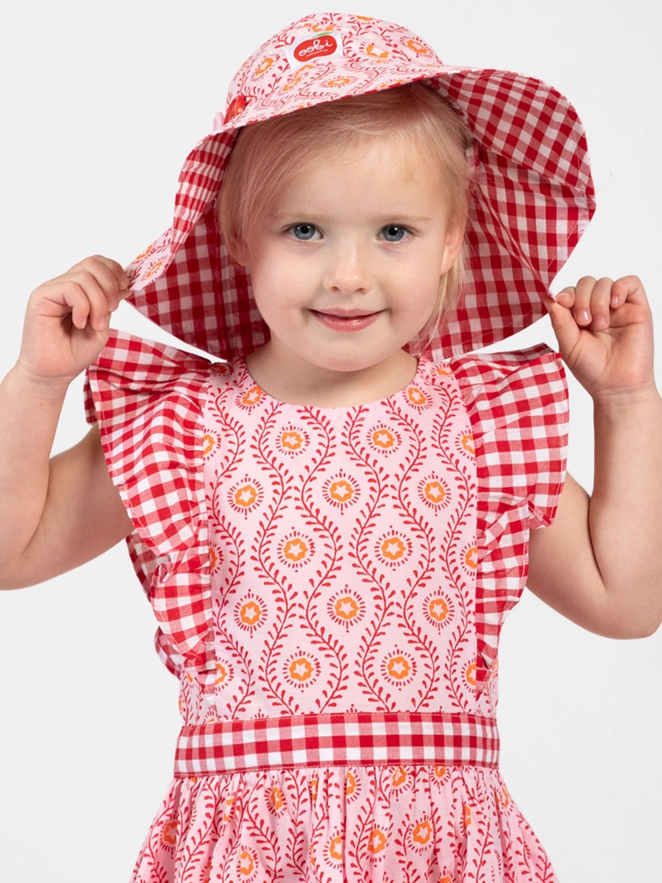 A young child smiles at the camera in the Woodblock Flower Jayne Dress by Woodblock Flower, paired with a matching wide-brimmed gingham hat, holding the hat with both hands against a plain background.