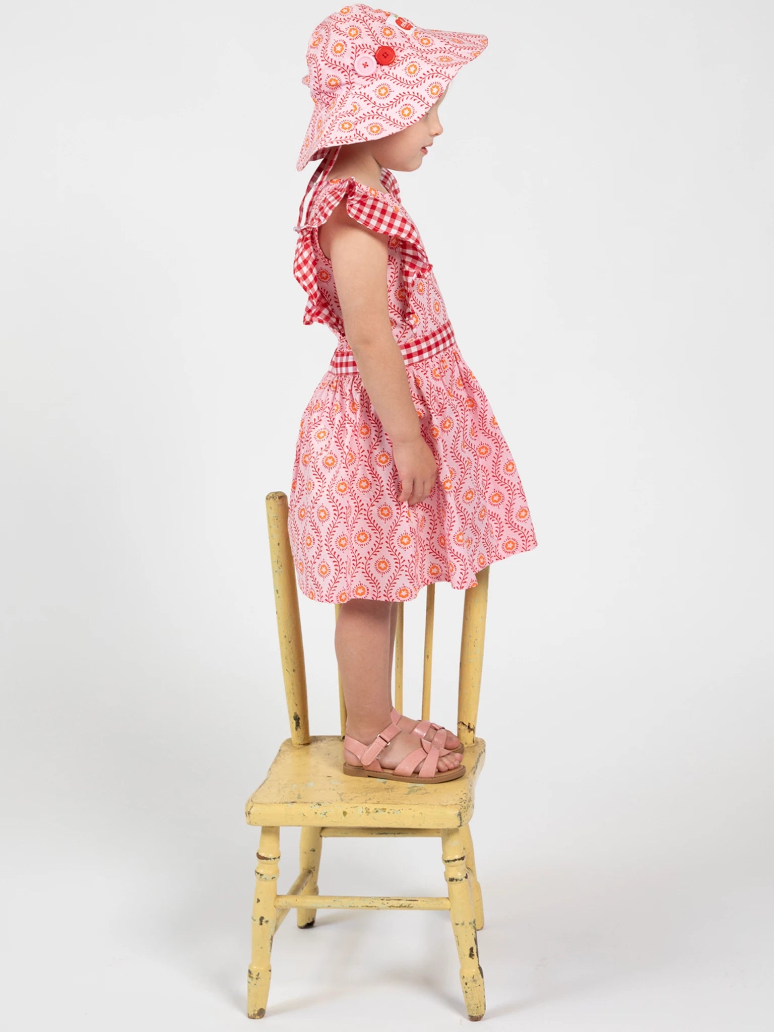 Child wearing a pink dress and hat sitting on a wooden chair against a white background
