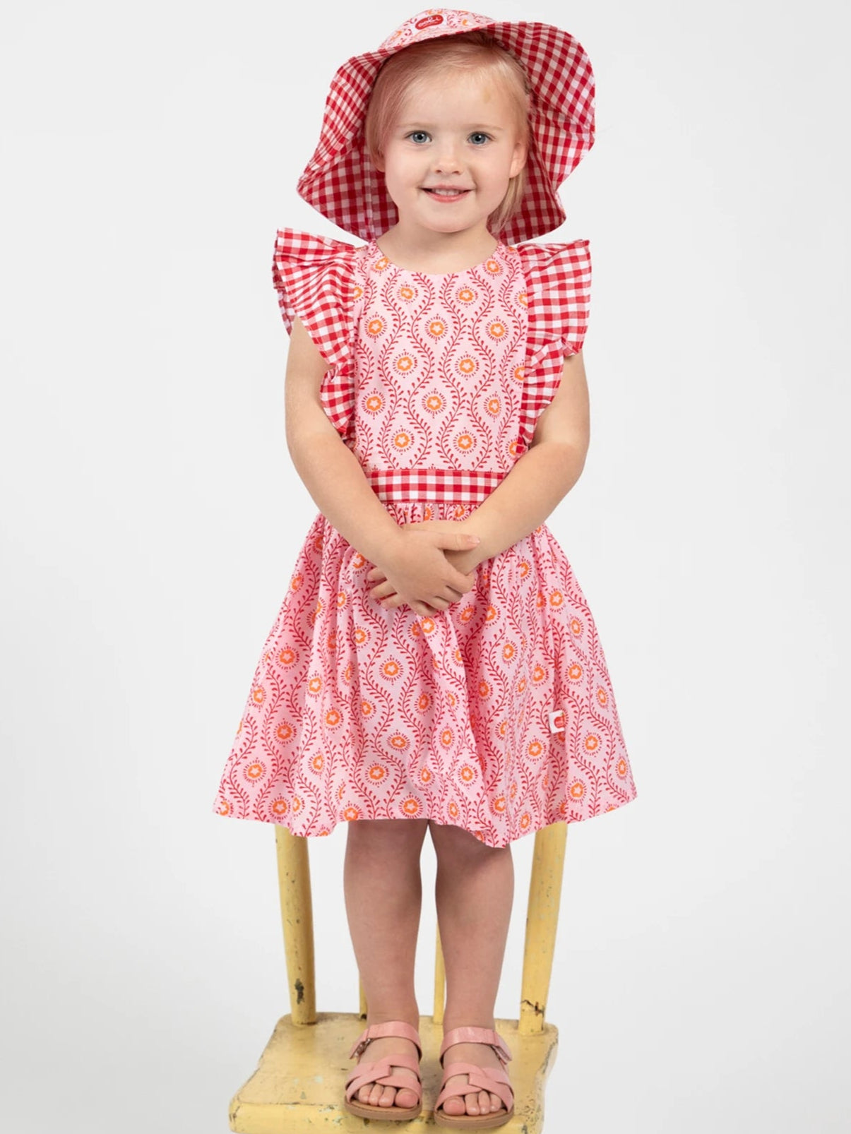 Young girl in a red and white checkered dress with a matching hat sitting on a wooden chair against a white background.