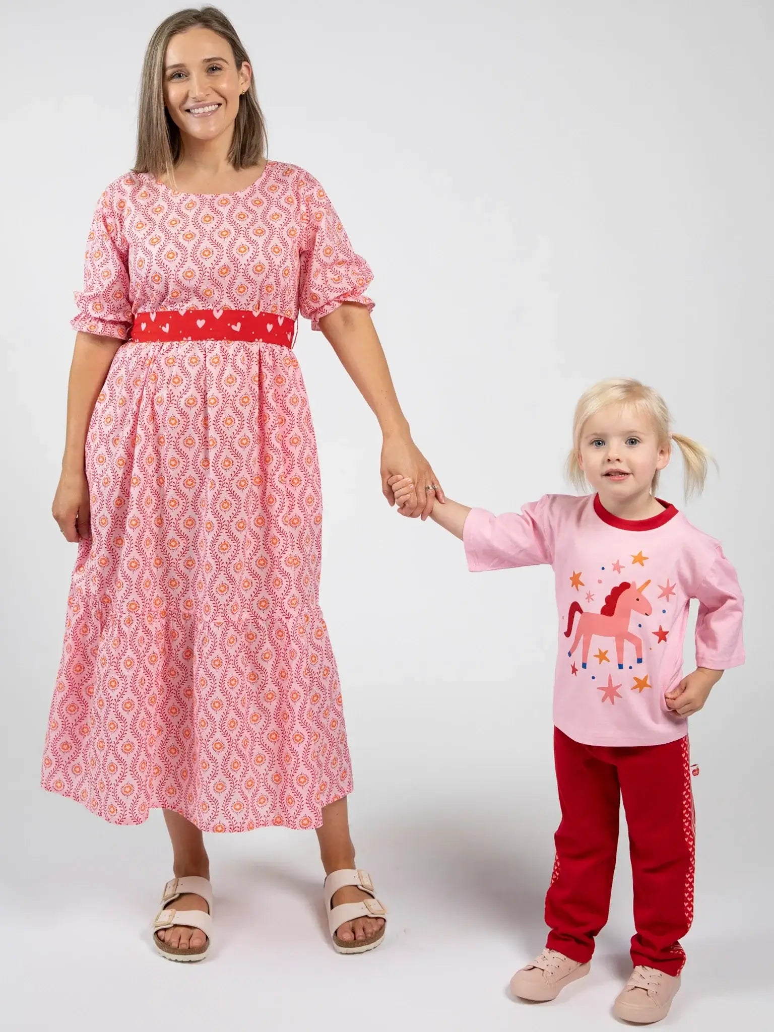 An adult in a Blue Stripe Women's Woodblock Maxi Dress stands in sandals, holding hands with a child in a pink horse-print shirt and red pants.