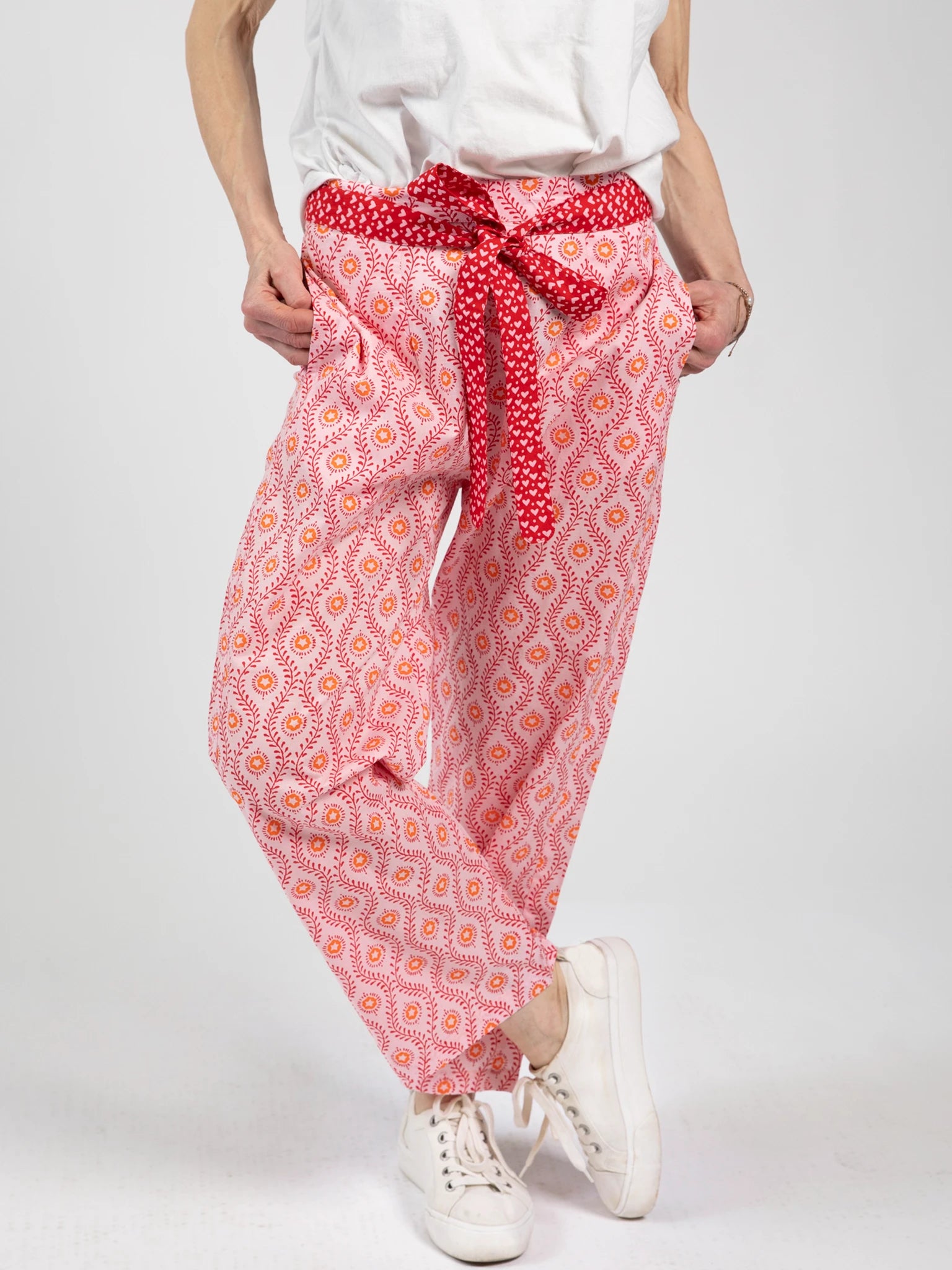 A person wears Woodblock Flower’s Woodblock Women's Resort Pants—loose pink patterned pants with a red sash—paired with white sneakers and a white top, standing against a plain background.