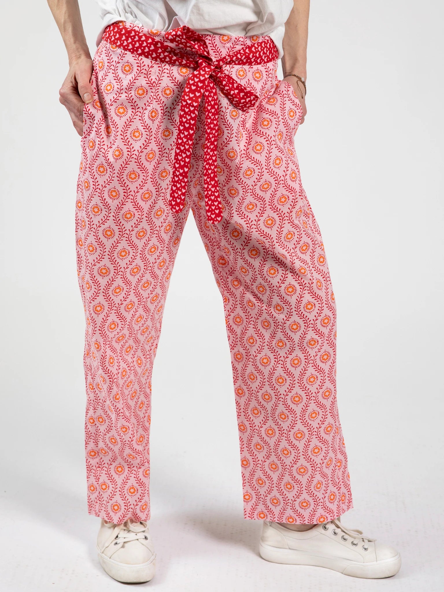 A woman models the Woodblock Flower Woodblock Women's Resort Pants—artisan-printed in pink and red with a red tie belt—paired with white sneakers and a white shirt, standing against a plain white background.