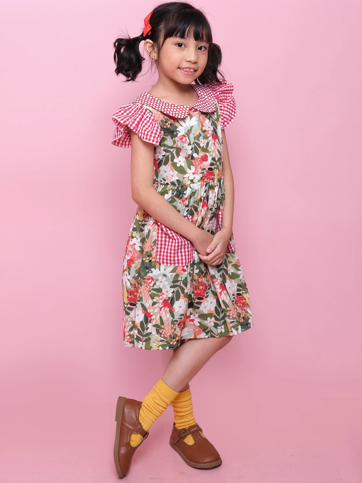 A young girl stands against a pink background, wearing the Aussie Bush Flowers Collared Dress by Aussie Bush Flowers, featuring 100% cotton floral fabric, red checkered accents, yellow socks, brown shoes, and a red hair bow.