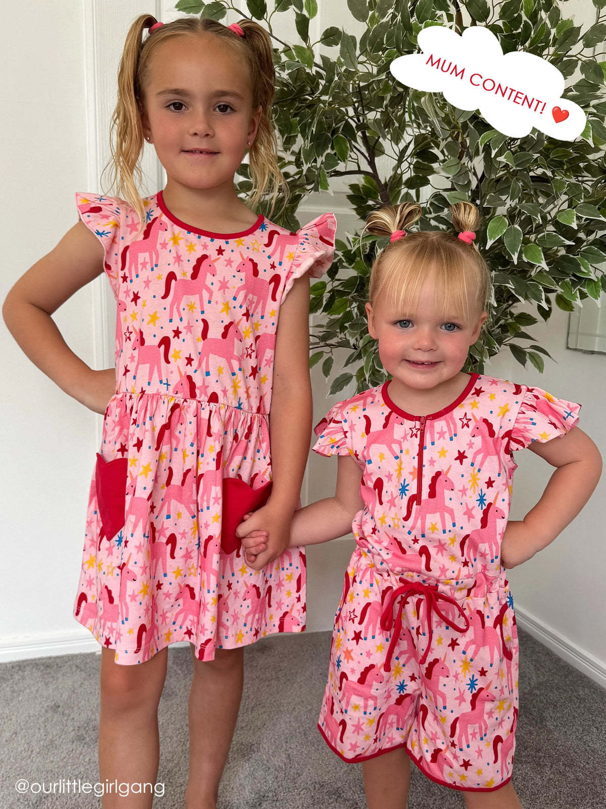 Two young girls standing indoors holding hands, posing and smiling for the camera in front of a potted plant. They are wearing matching pink outfits patterned with pink and red horses, yellow stars, and blue sparkles.