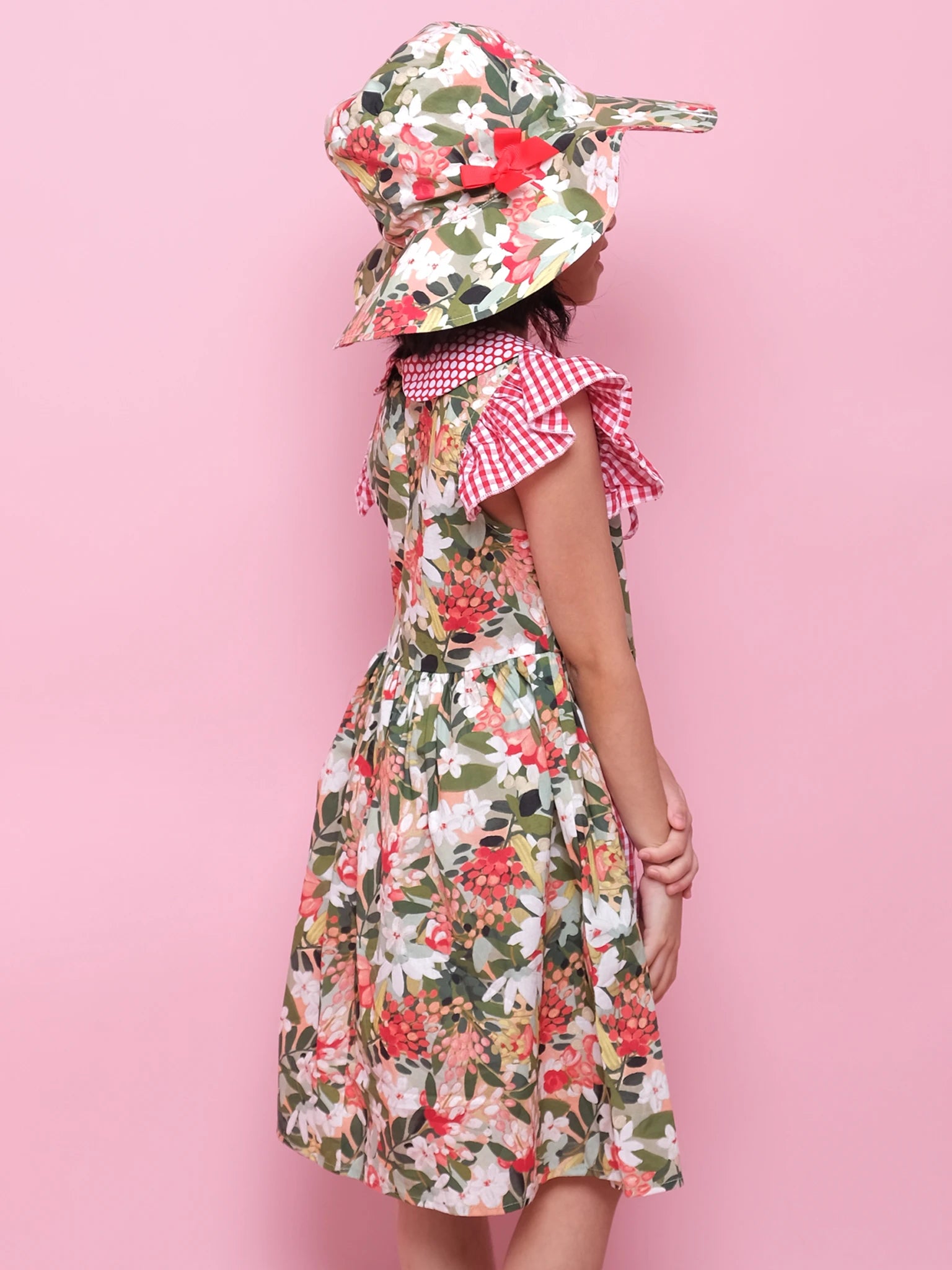 A child stands sideways against a pink background, wearing the Aussie Bush Flowers Ponytail Hat and a floral dress with a red and white gingham collar.